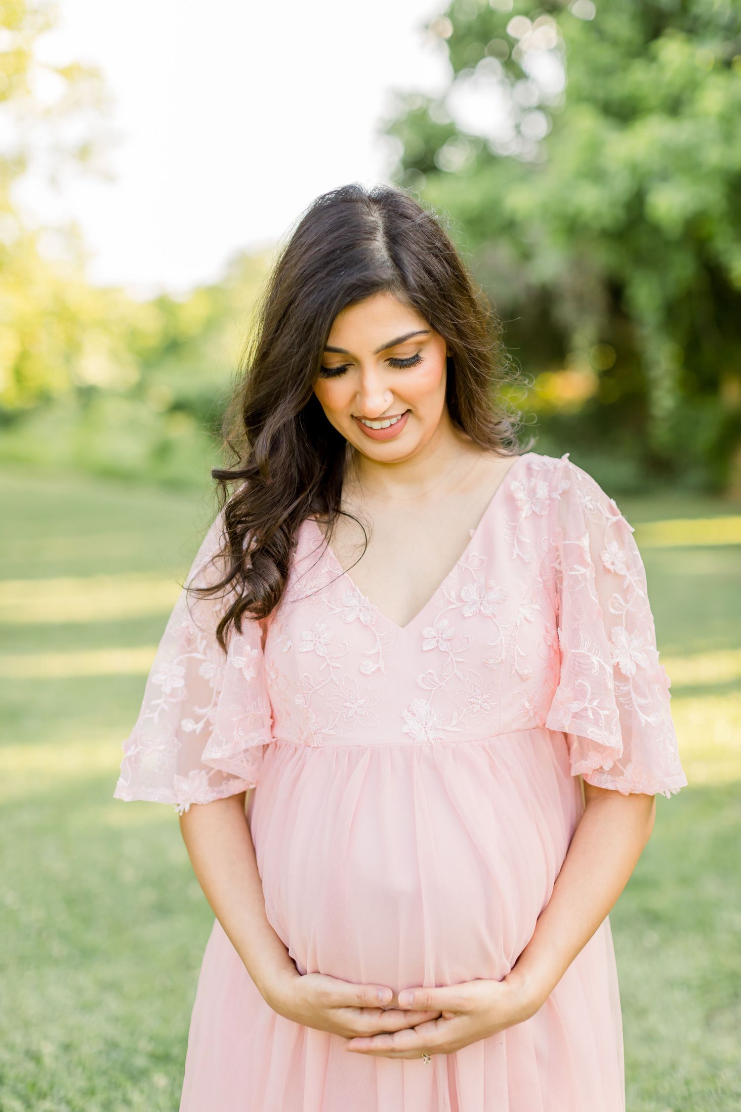 Woman in pink lace dress smiles while holding her baby bump in outdoor maternity photo.