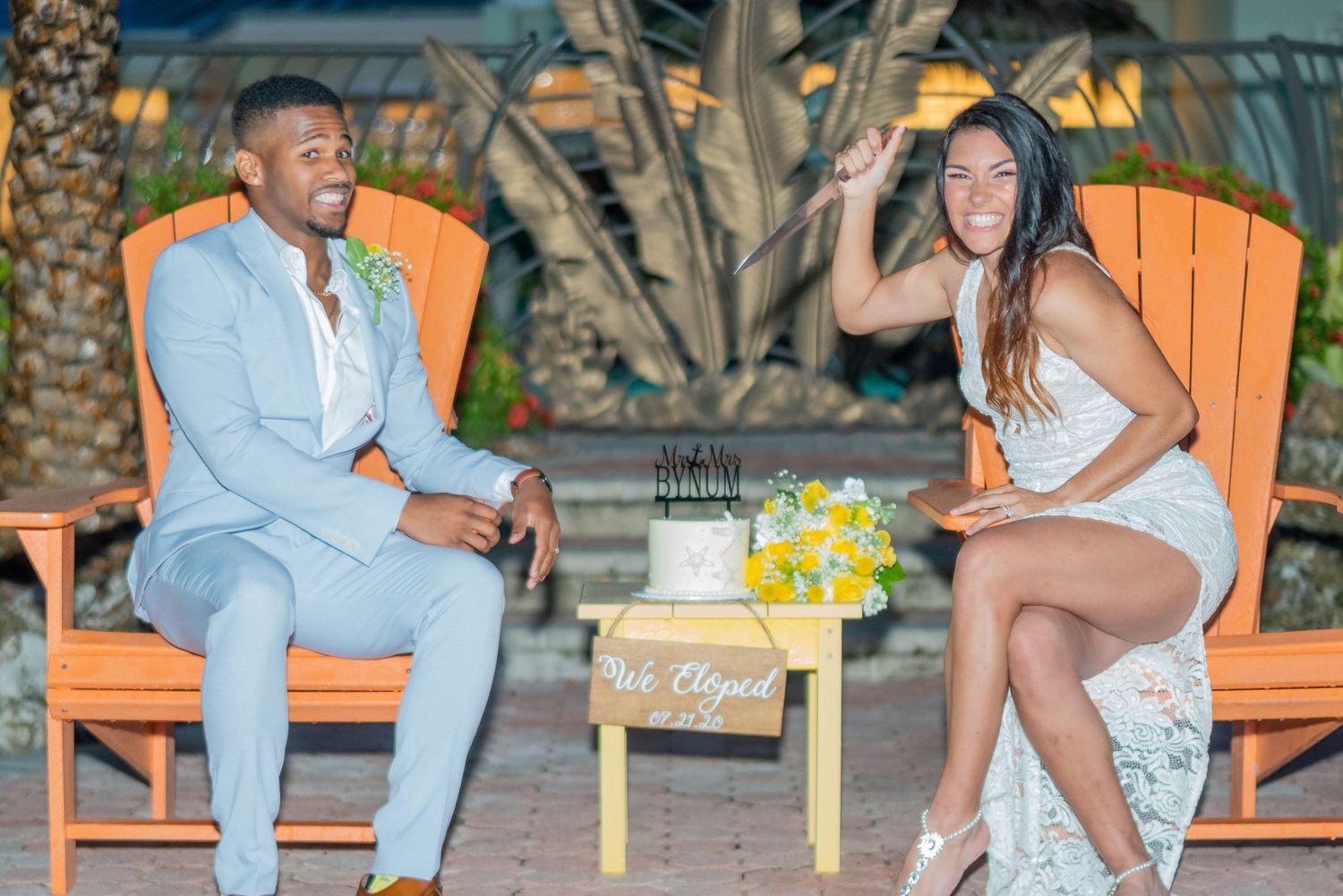 Couple in wedding attire sitting in orange Adirondack chairs with yellow flowers on a small table between them.