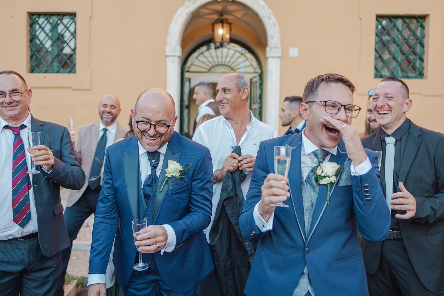 Groomsmen in navy suits raising glasses in celebratory toast outside church.