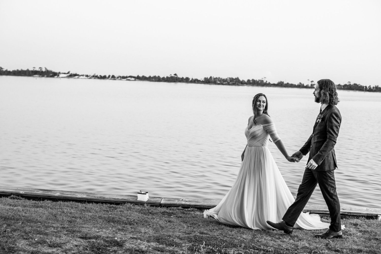 A black and white photo of a couple walking hand in hand by a serene lakeside.