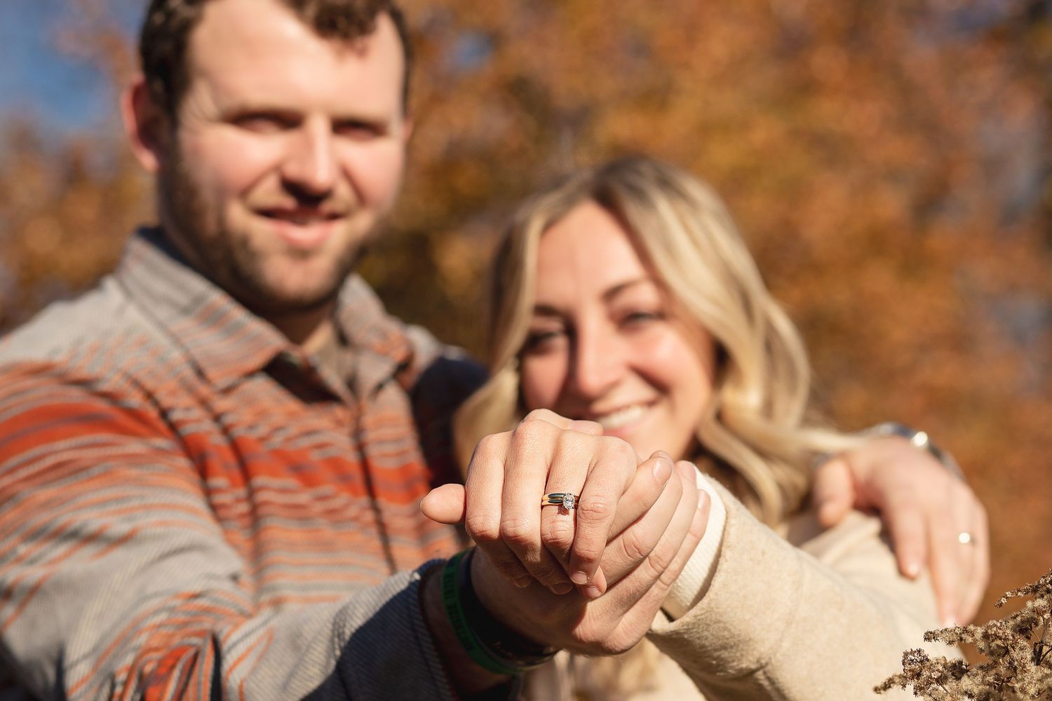 Couple showing off engagement ring in sunny autumn setting with fall foliage.