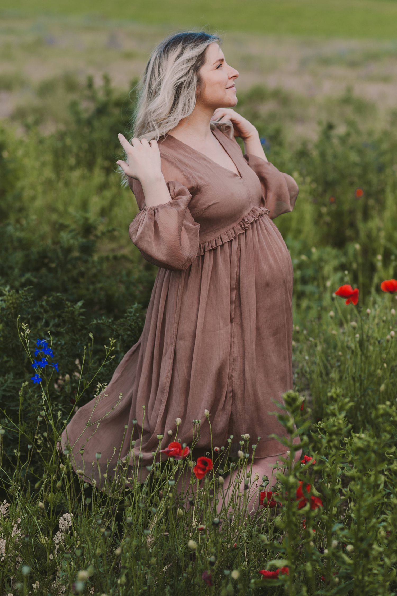 Woman in mauve dress standing among red poppies and wildflowers in a natural field setting.
