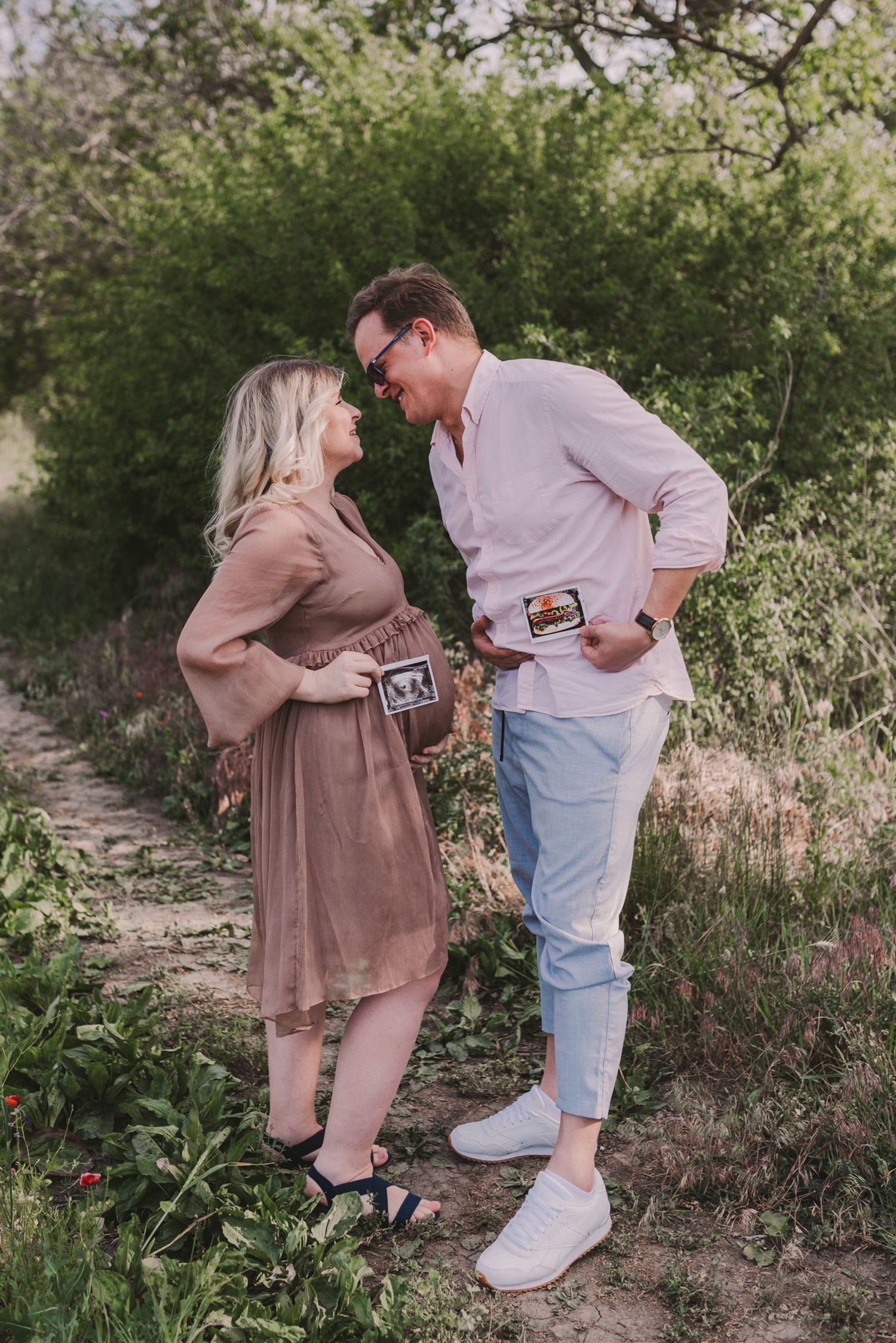 Couple sharing a playful moment while looking at ultrasound photos in a garden setting.