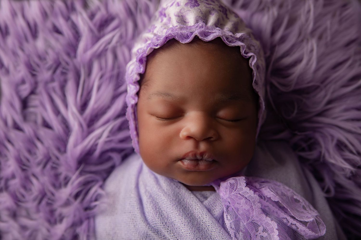Sleeping newborn in purple bonnet and wrap against soft purple textured background.