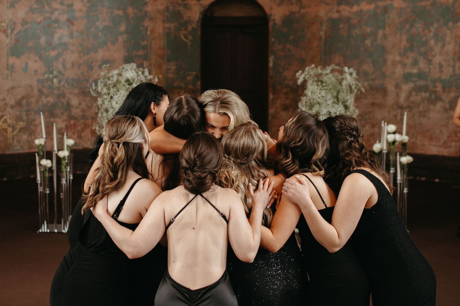 Bridesmaids in black dresses huddle together in a circular group hug at a rustic venue.