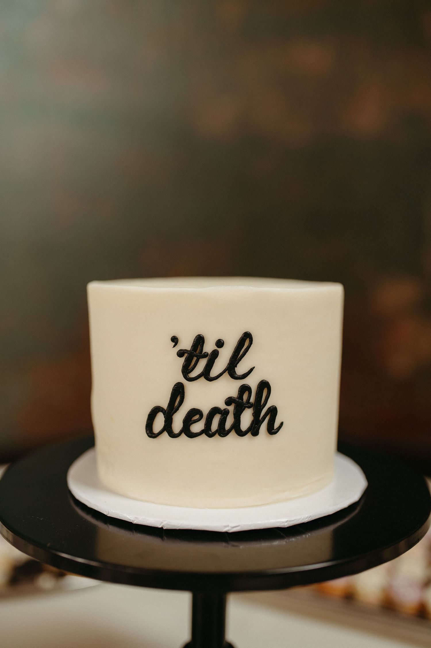 A minimalist white wedding cake with 'til death' written in black script lettering on a black cake stand.