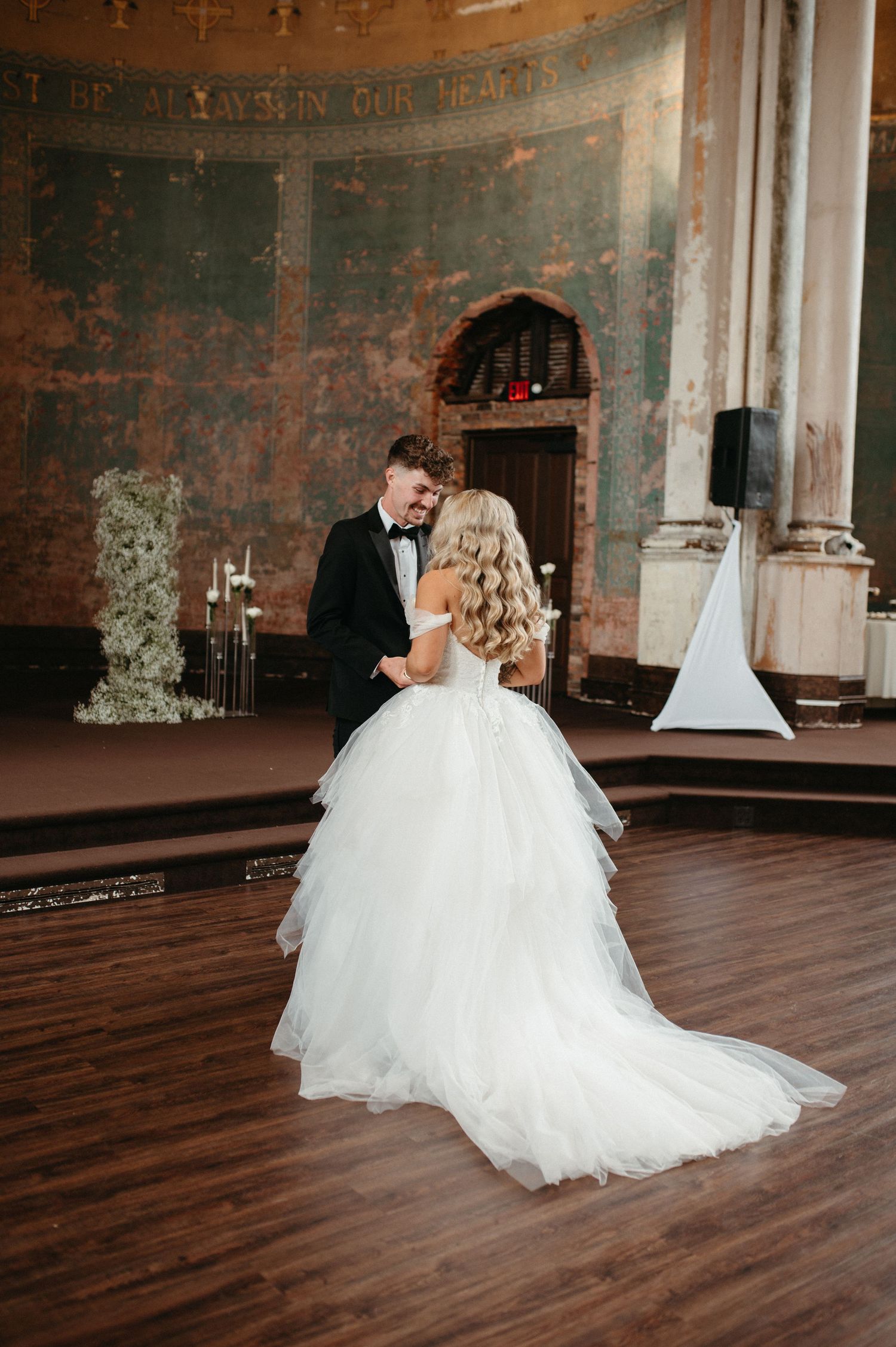 Couple dances in elegant venue with rustic walls and hardwood floors.
