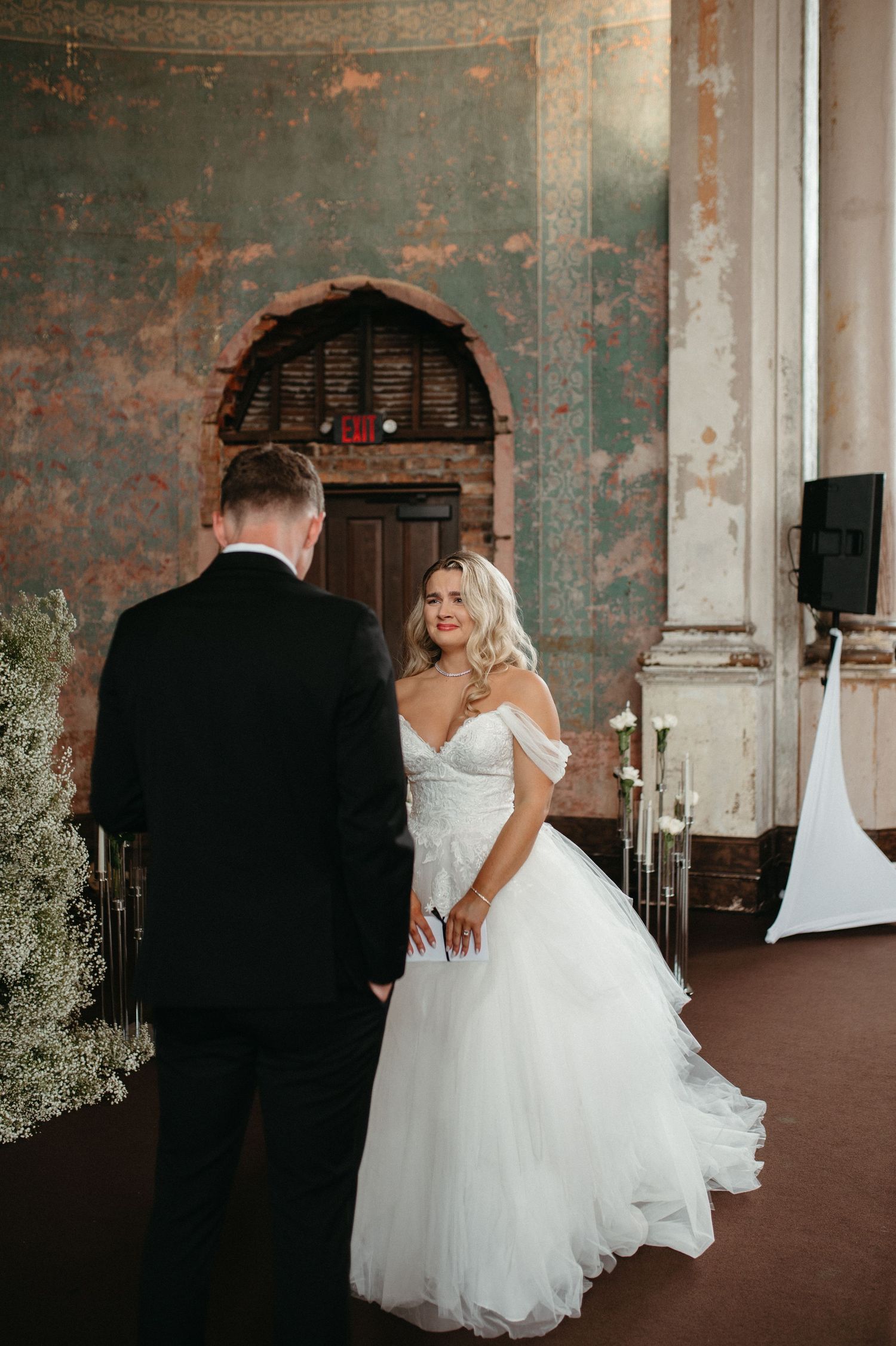 A wedding first look moment in an elegant venue with distressed turquoise walls and floral arrangements.