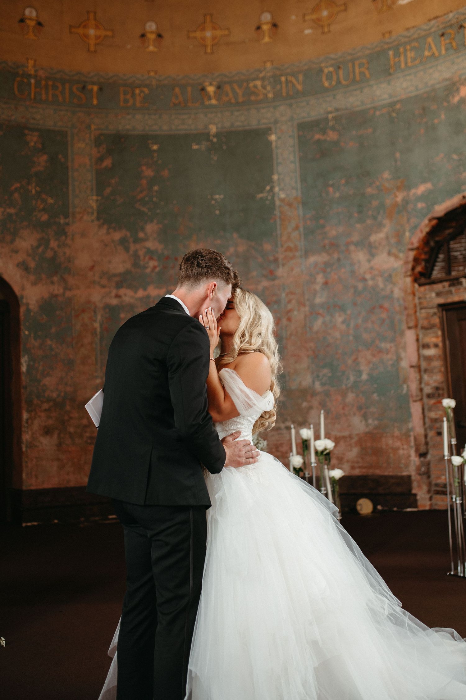 An intimate wedding ceremony moment against weathered walls with religious text visible above.