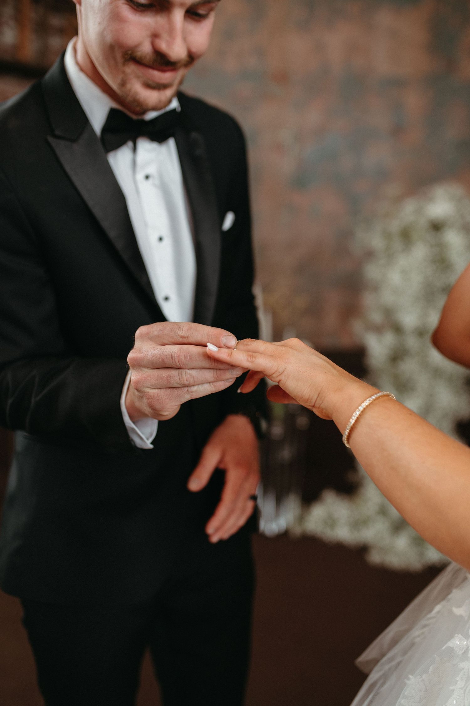 Close-up of wedding ring exchange during ceremony with classic black tuxedo details.