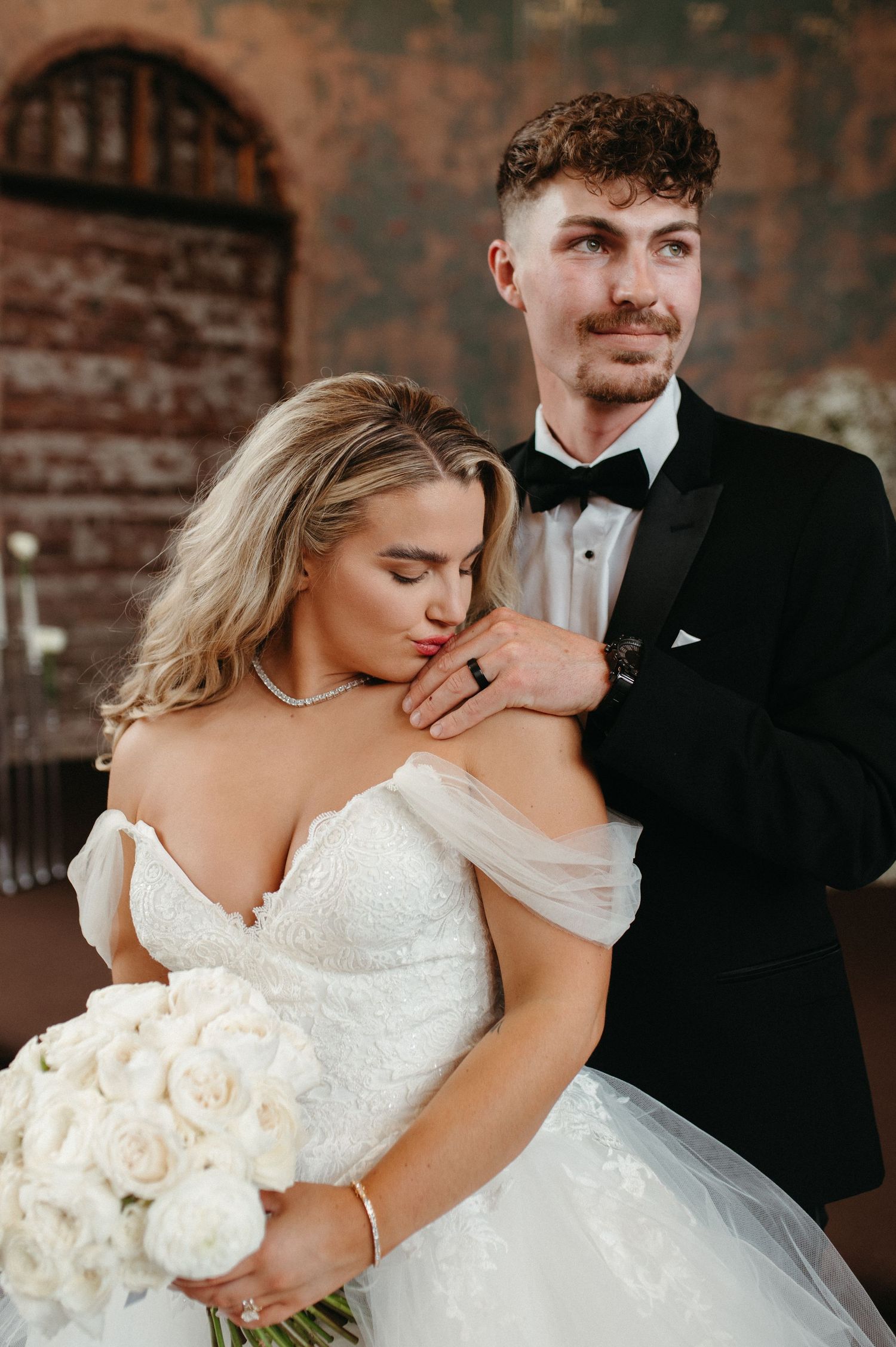 Romantic wedding portrait with bride holding white rose bouquet against rustic brick backdrop.