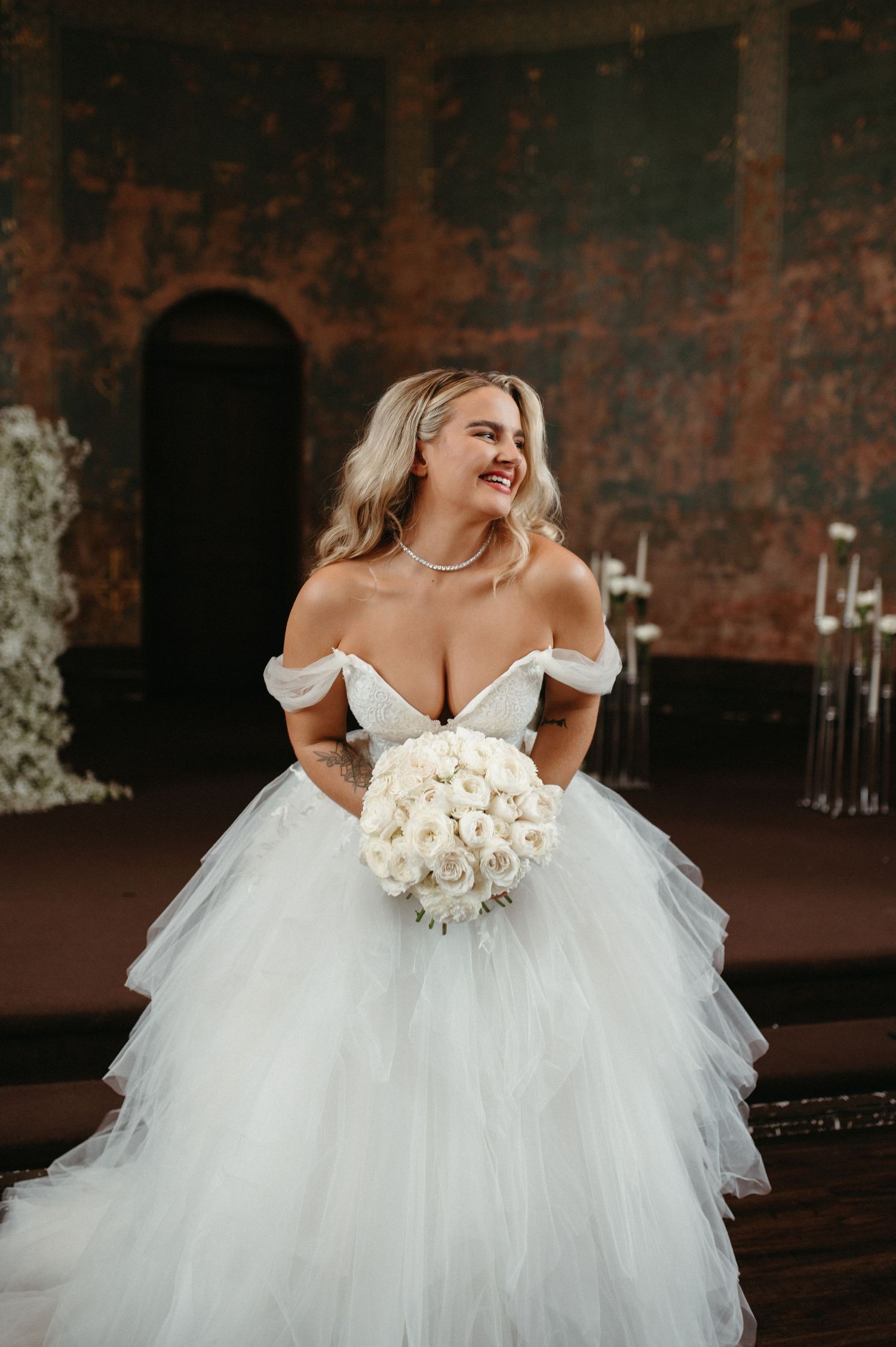 Bride in a stunning off-shoulder ball gown holding white roses against a rustic backdrop.