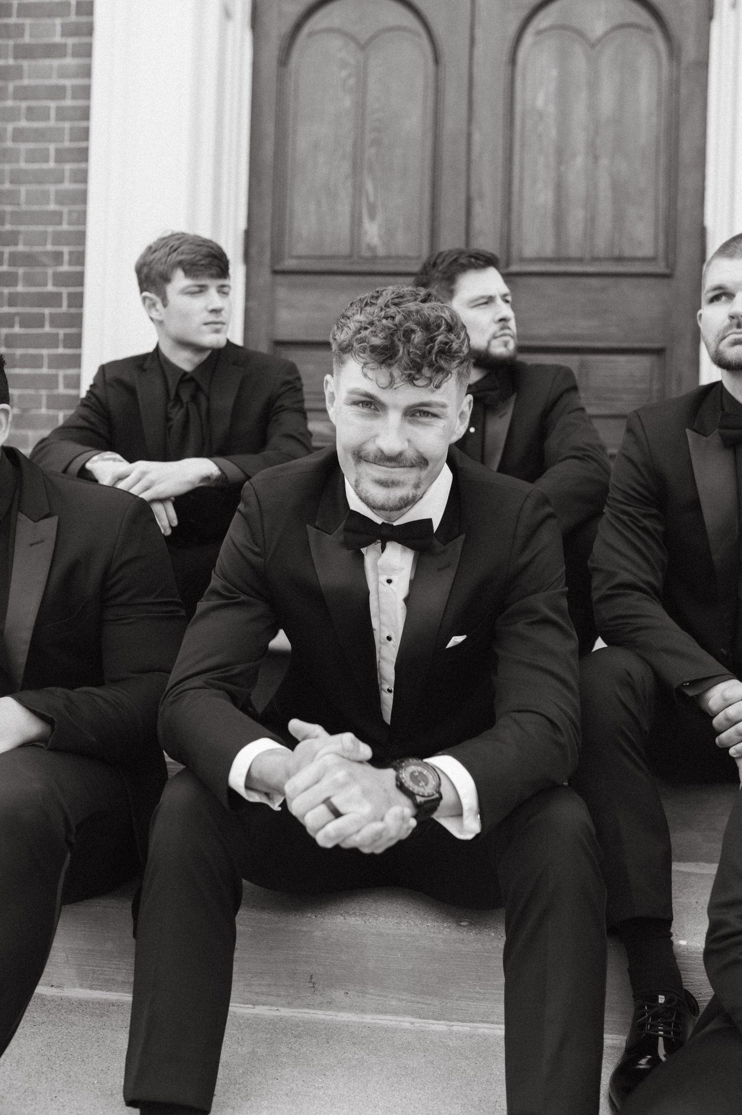 Group of groomsmen in formal black tuxedos sitting on church steps.