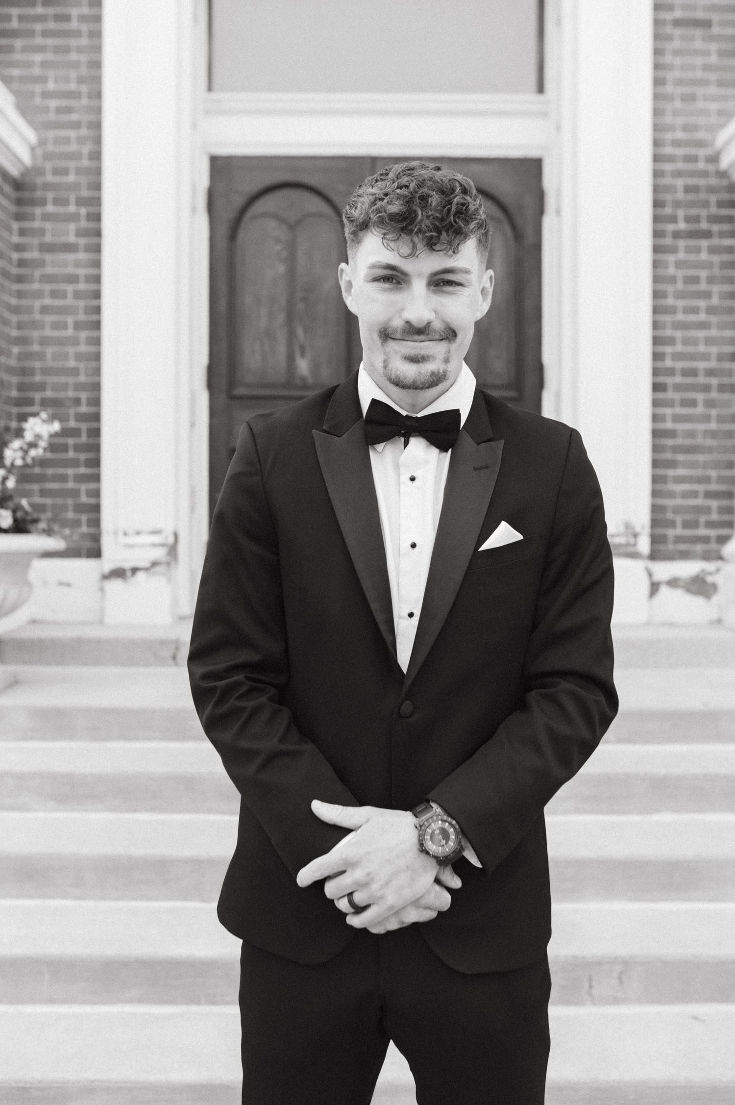 Groom in classic black tuxedo standing on church steps.