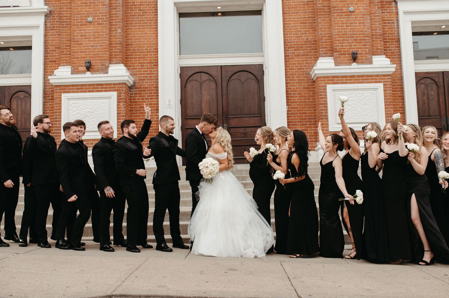 Wedding party in black attire stands with couple in front of brick church entrance celebrating with raised arms.
