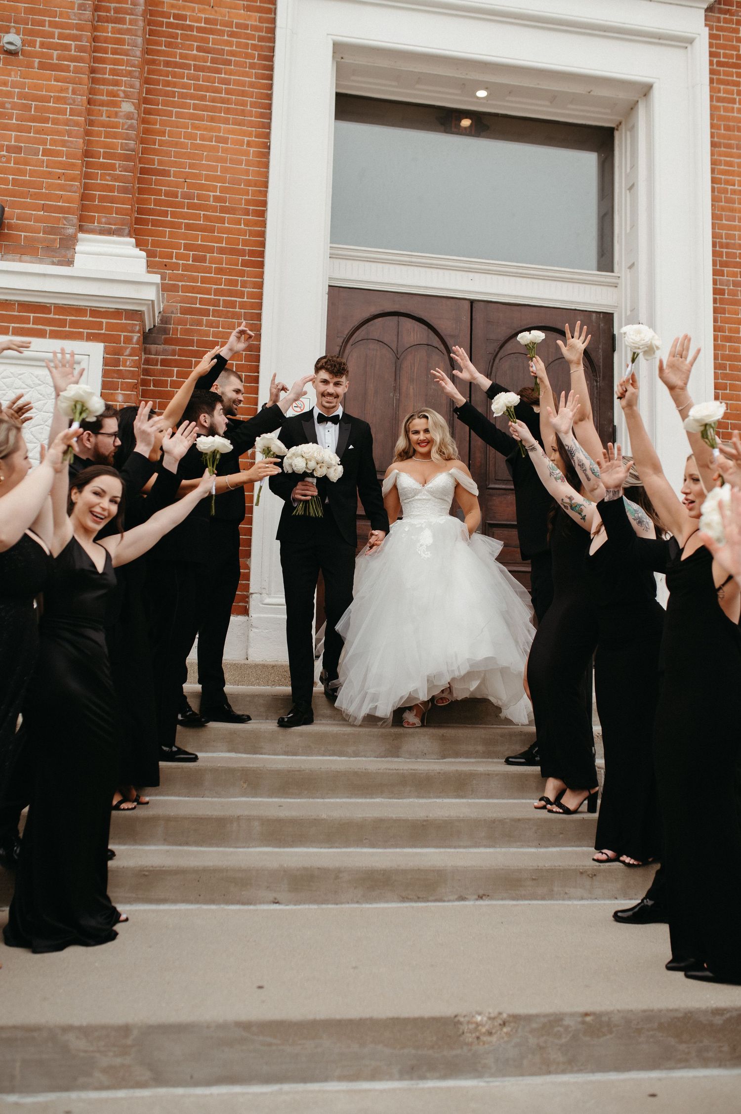 Wedding party cheers as newlyweds descend church steps with bride in flowing white gown.