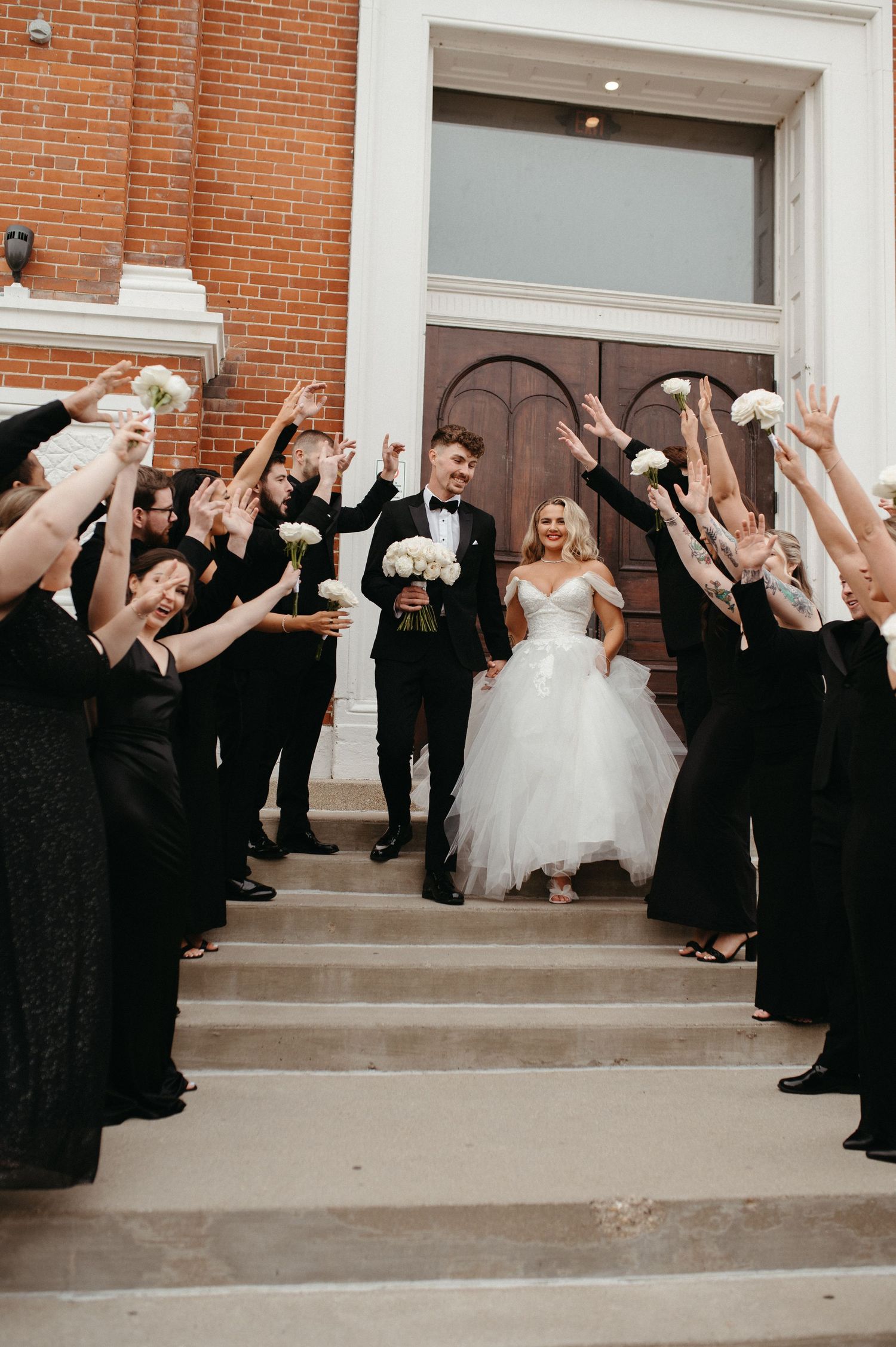 Bridal party in black dresses and suits forms aisle on church steps celebrating the newlyweds.
