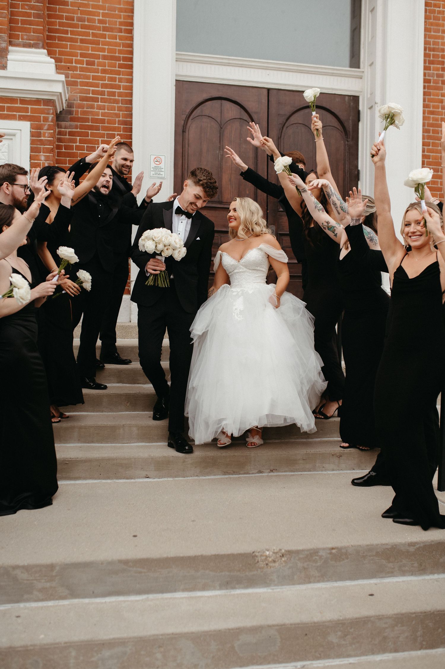 Bridal party in elegant black attire cheers as couple celebrates on church entrance steps.