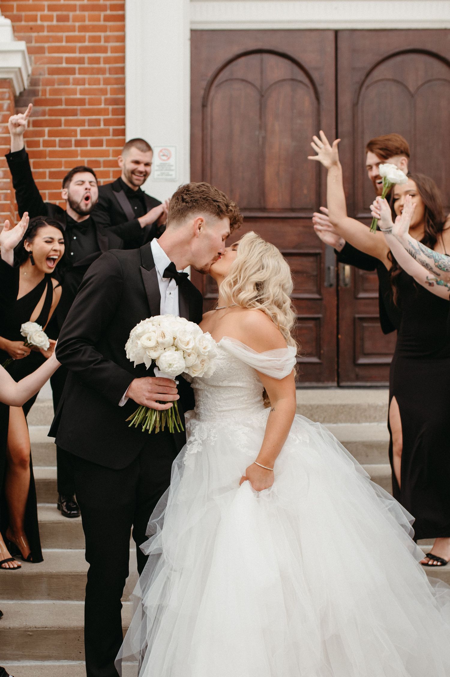 Joyful wedding party celebrates as couple shares romantic moment on church steps.
