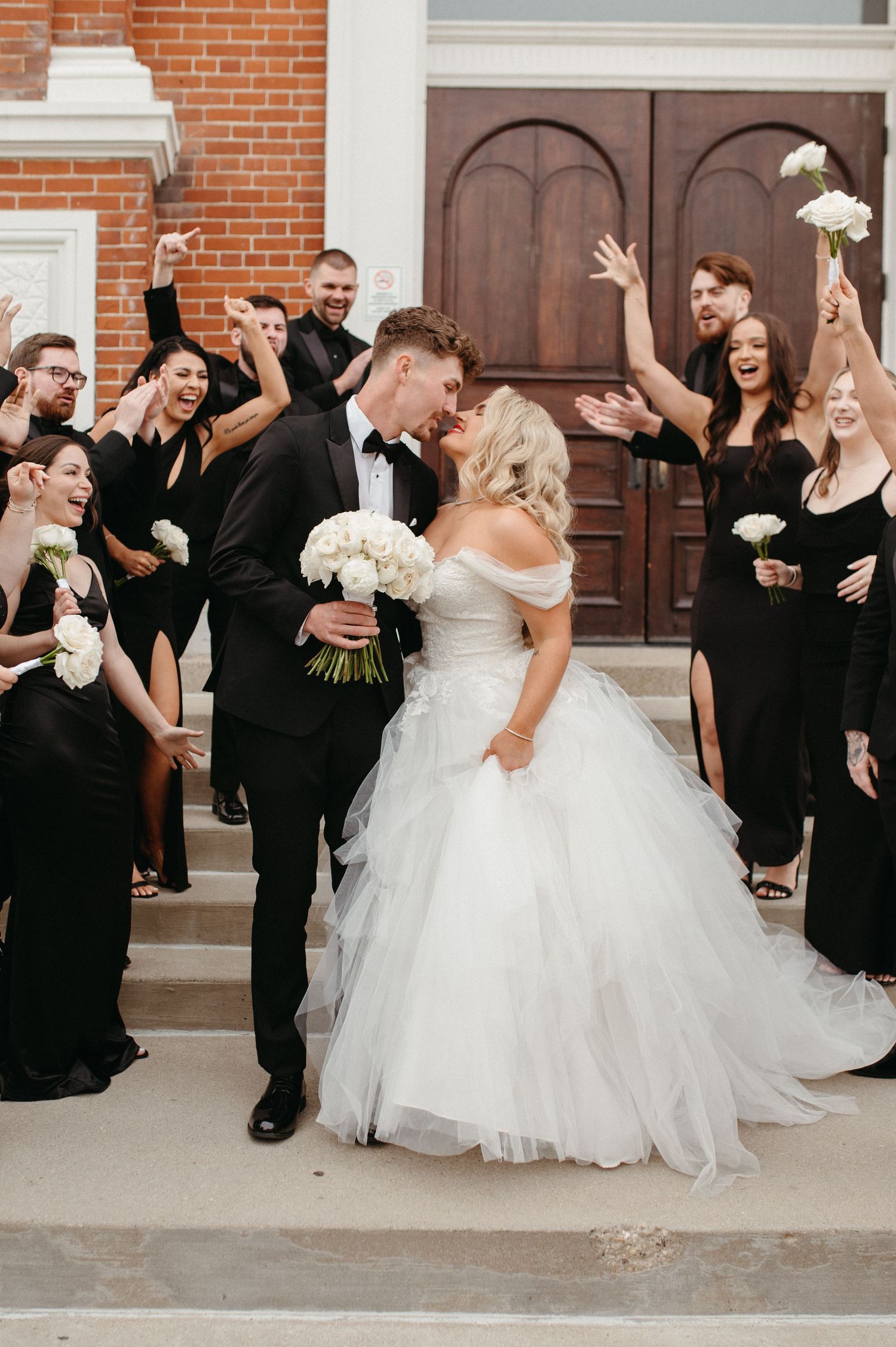 Wedding party raises arms in celebration as couple shares intimate moment on church steps.