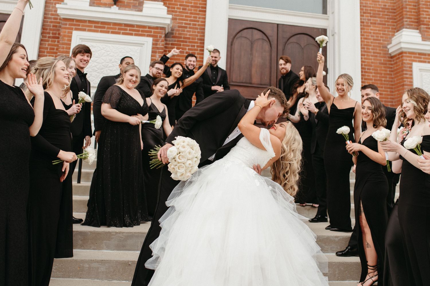 Wedding party in black attire celebrates with couple during romantic moment on church steps.