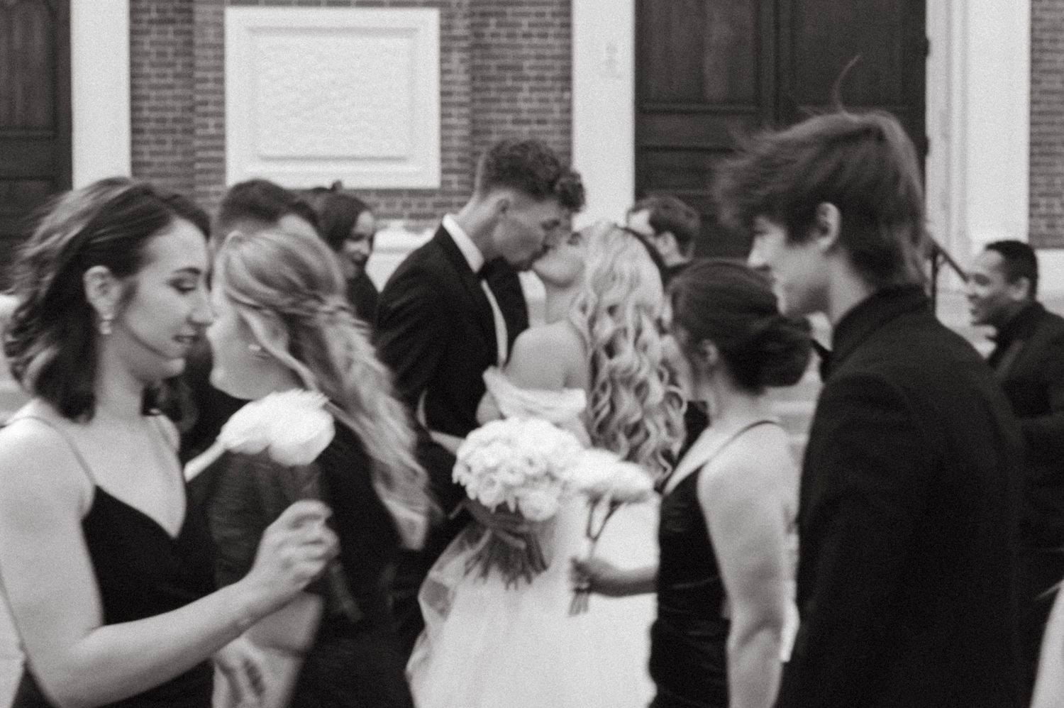 Black and white photo captures intimate wedding party moment outside historic brick church.