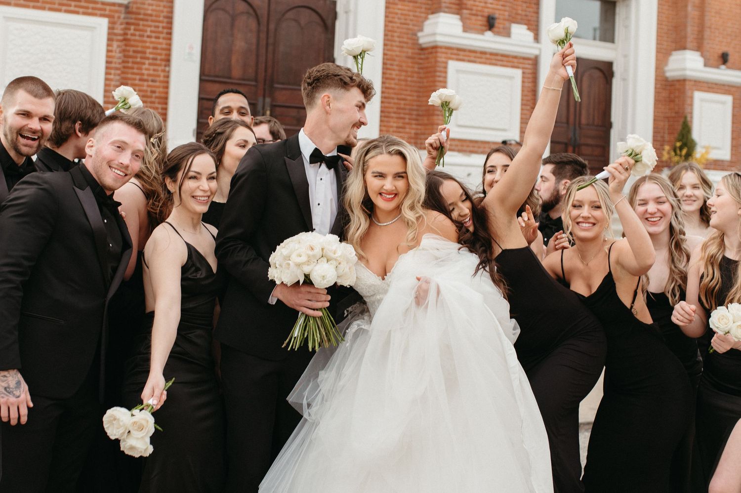 Wedding party celebrates on church steps with bride in white gown surrounded by attendants in black.