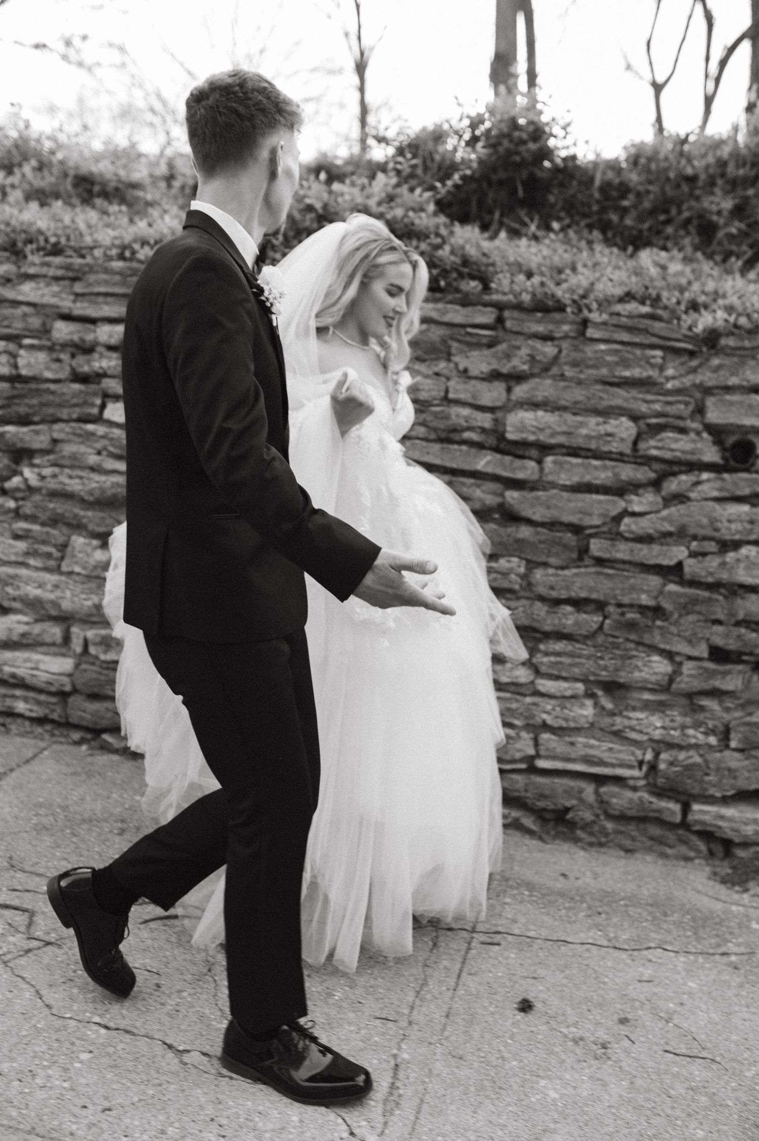 Black and white image of couple walking along stone wall path in wedding attire.