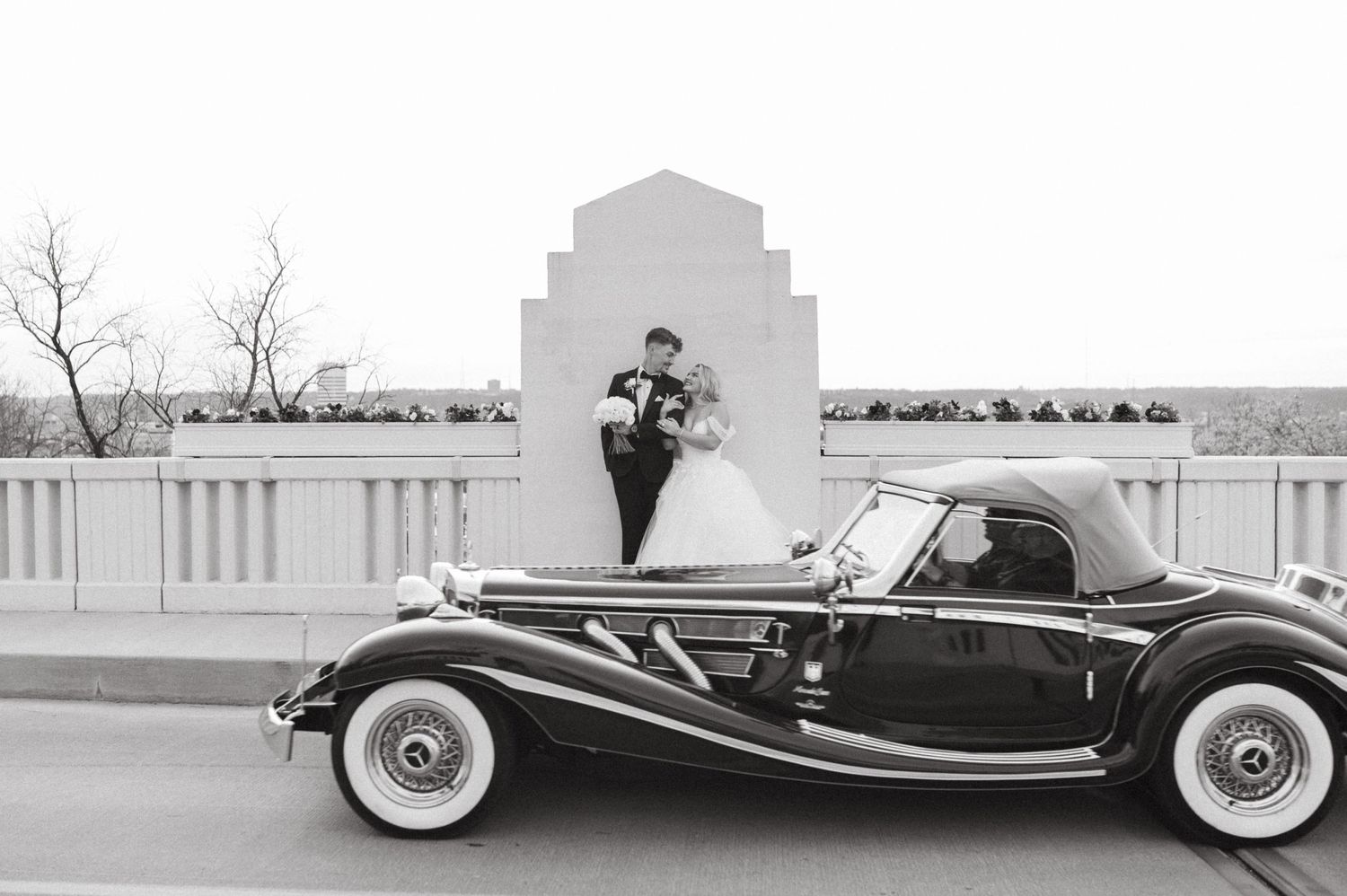 Classic vintage car parked beside white architectural structure in elegant black and white wedding photo.