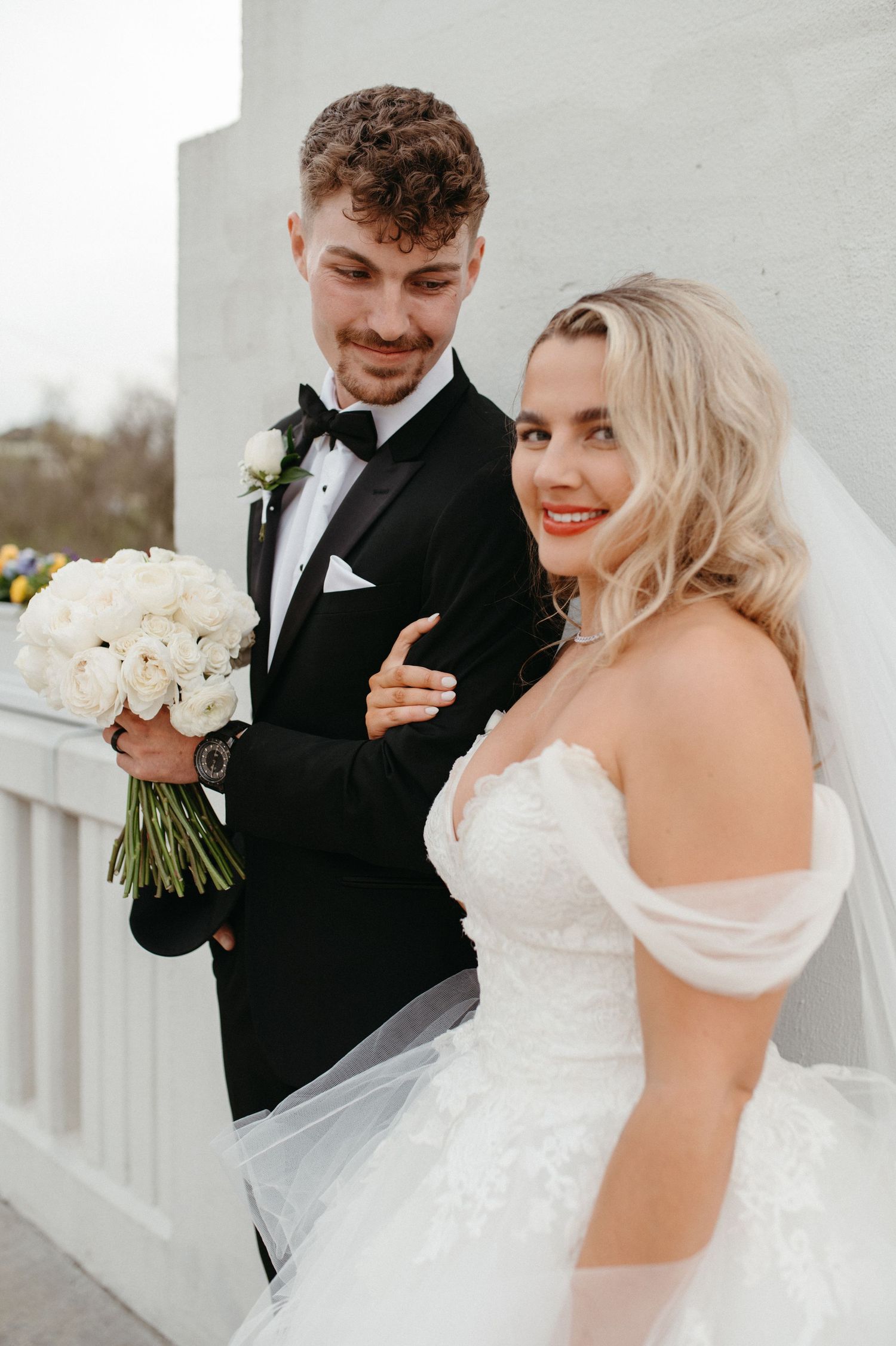 Elegant bride and groom pose with white bouquet against white pillar backdrop.