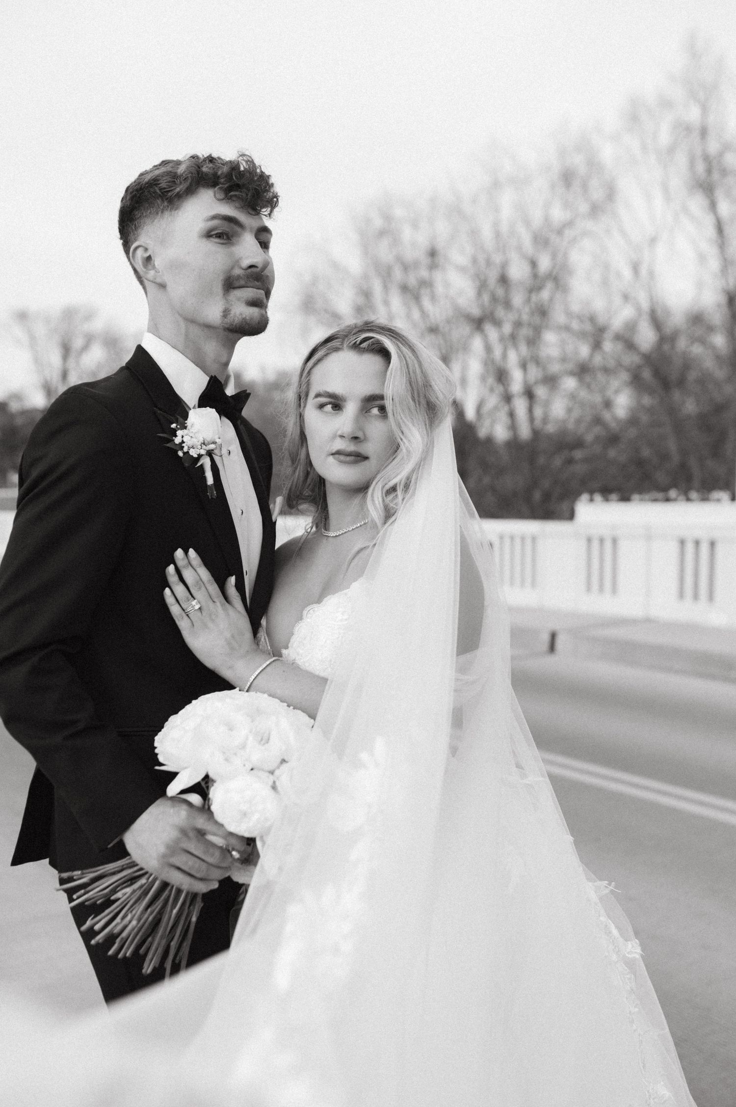 Black and white wedding portrait with couple holding bouquet against white railing.