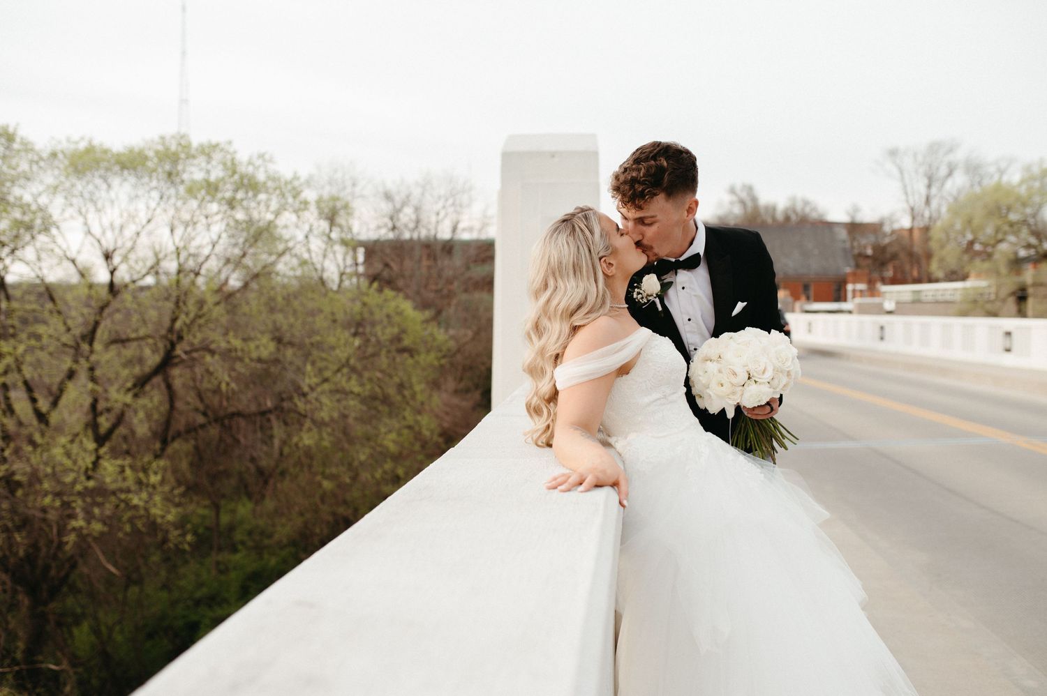 A newlywed couple embraces on a bridge while holding a white flower bouquet.