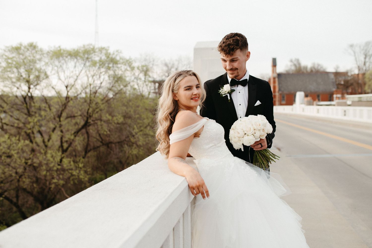 A bride in a flowing tulle gown leans against a white railing as her groom stands beside her.