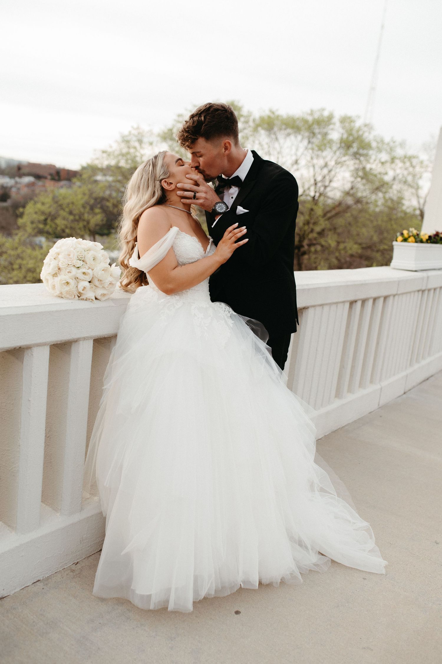 A romantic wedding embrace on a white bridge railing during golden hour.
