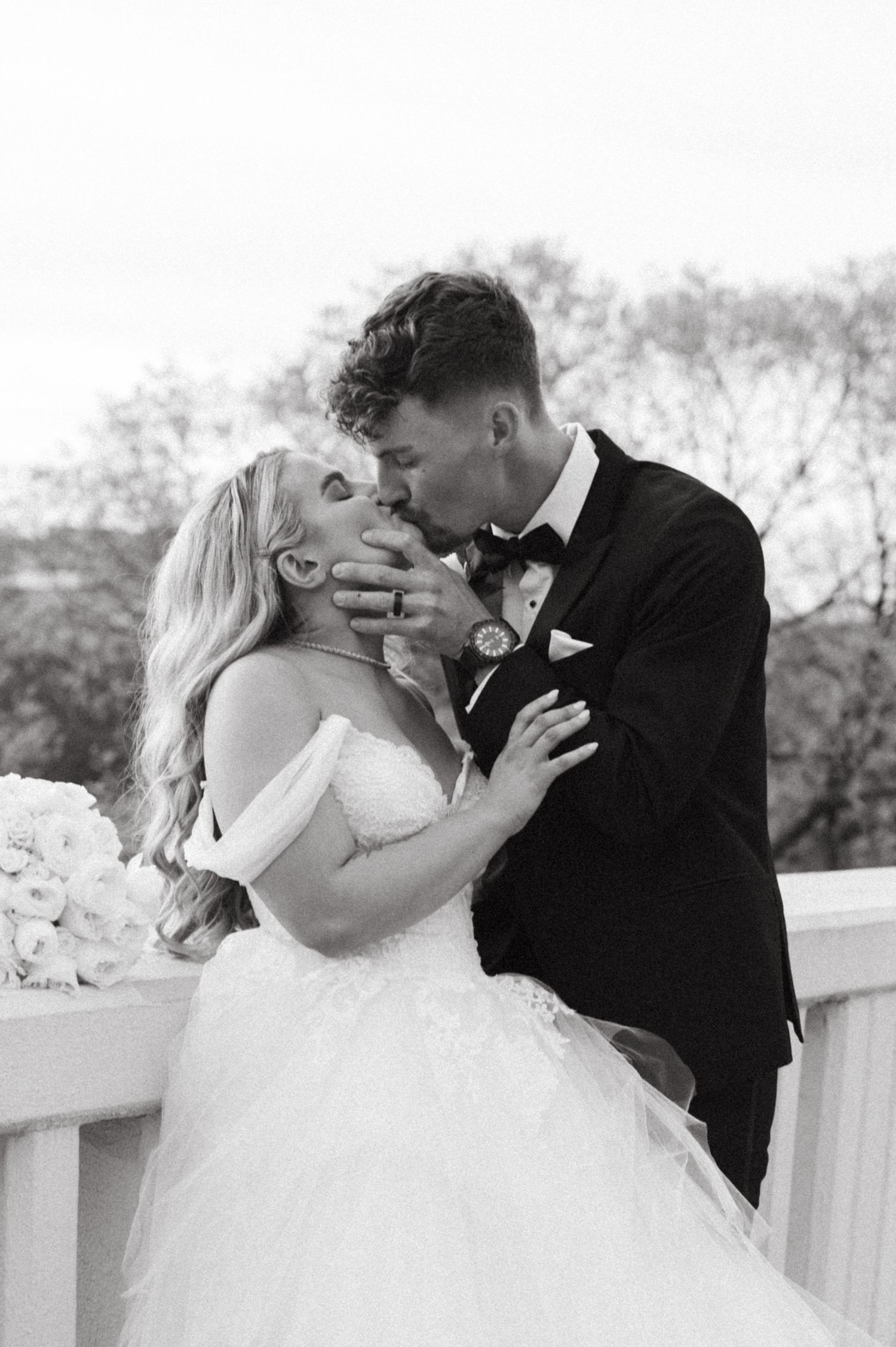A black and white wedding photo captures an intimate moment between newlyweds on a bridge.