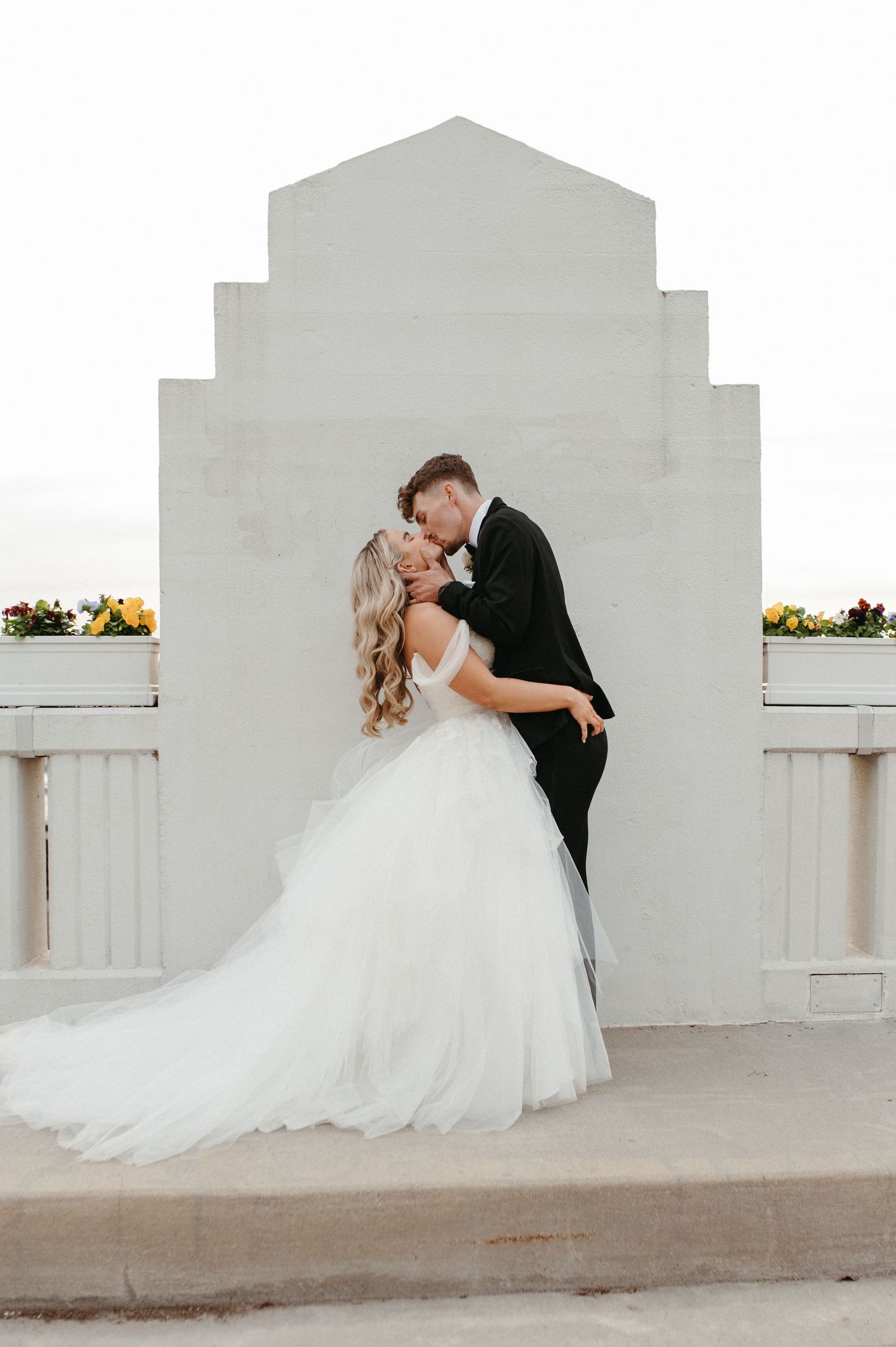 A romantic wedding couple shares a kiss against a white architectural backdrop with yellow flowers.