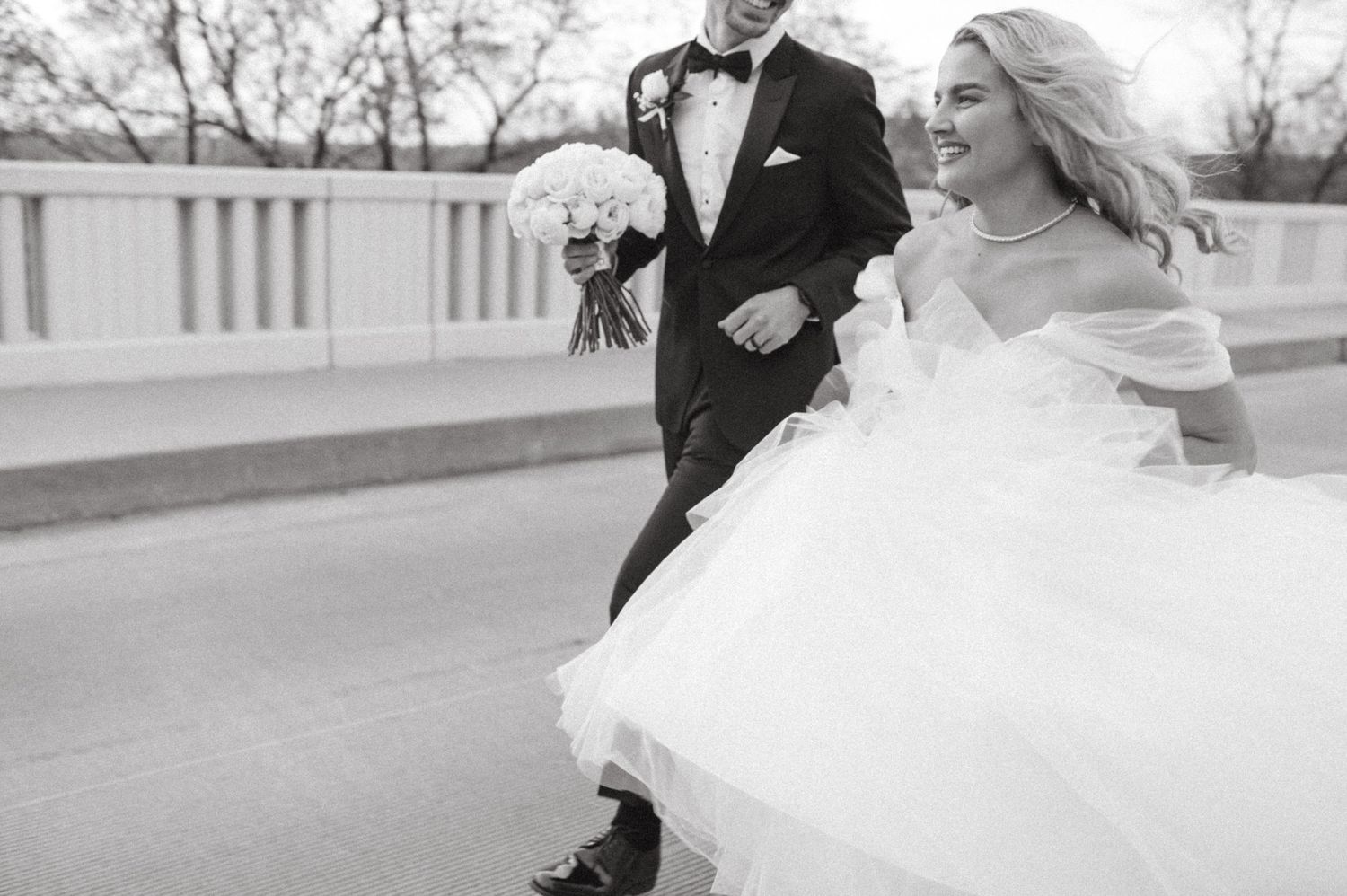 A joyful black and white wedding photo capturing movement and laughter on a bridge.