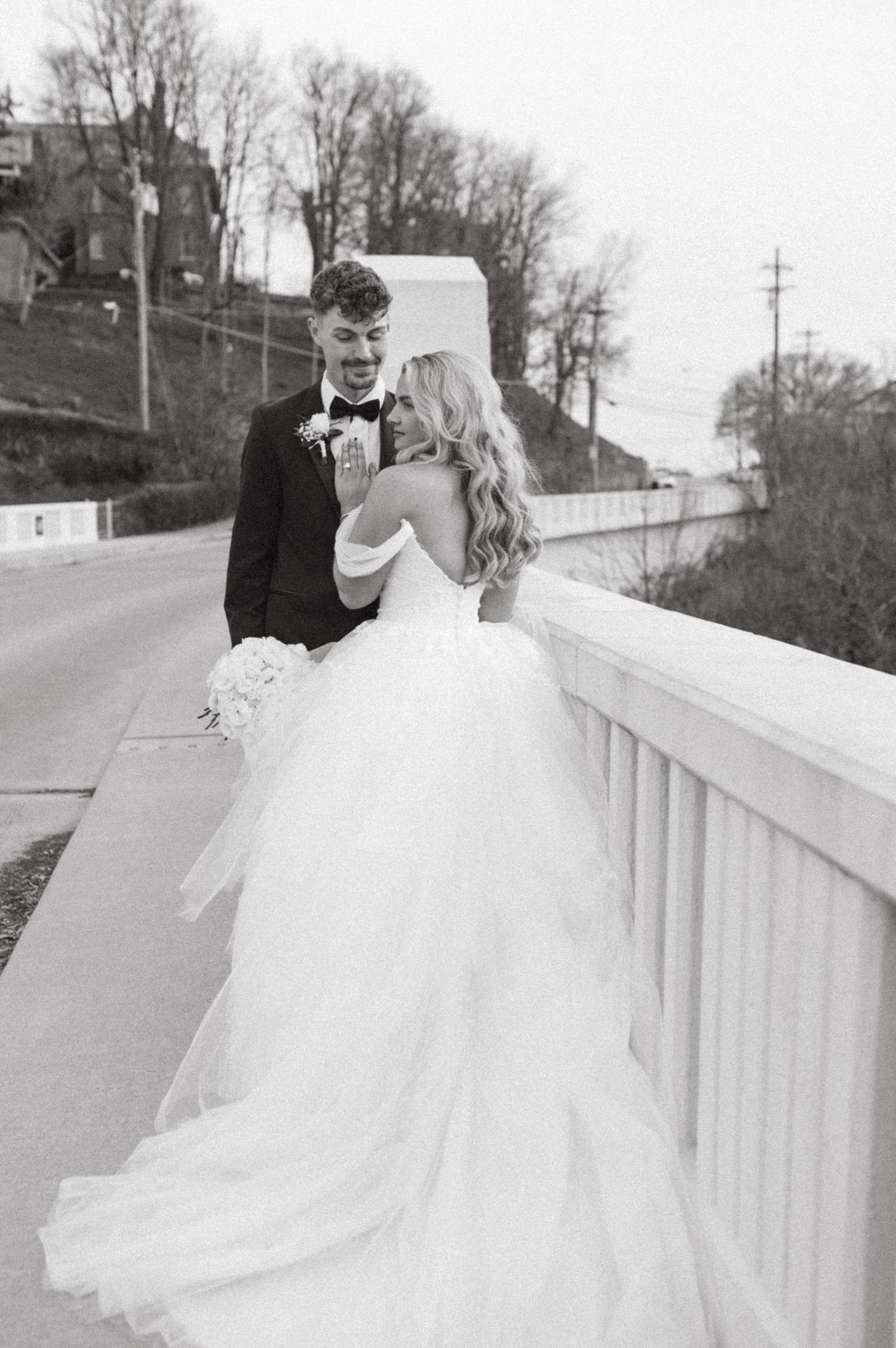 A candid wedding moment photographed in black and white on a bridge with bare trees in background.