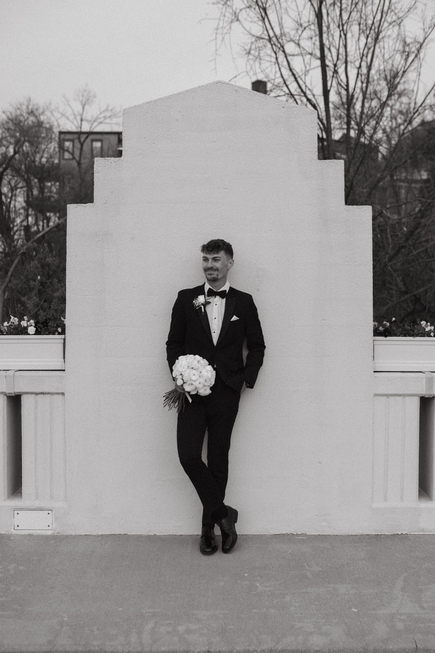 A formal black and white portrait of a groom holding a bouquet against a white architectural wall.