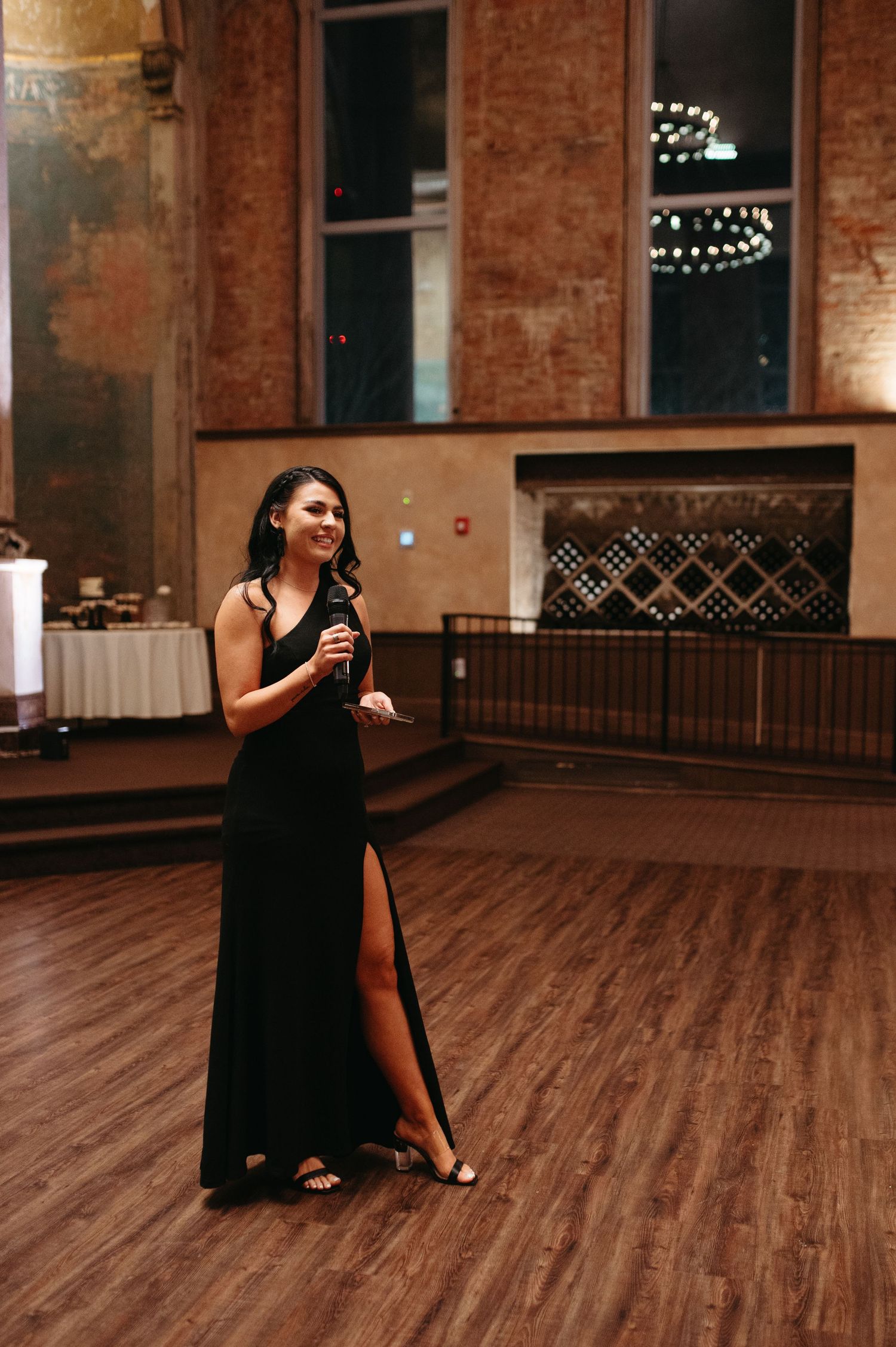 Woman in black formal dress giving speech on wooden dance floor at wedding reception.