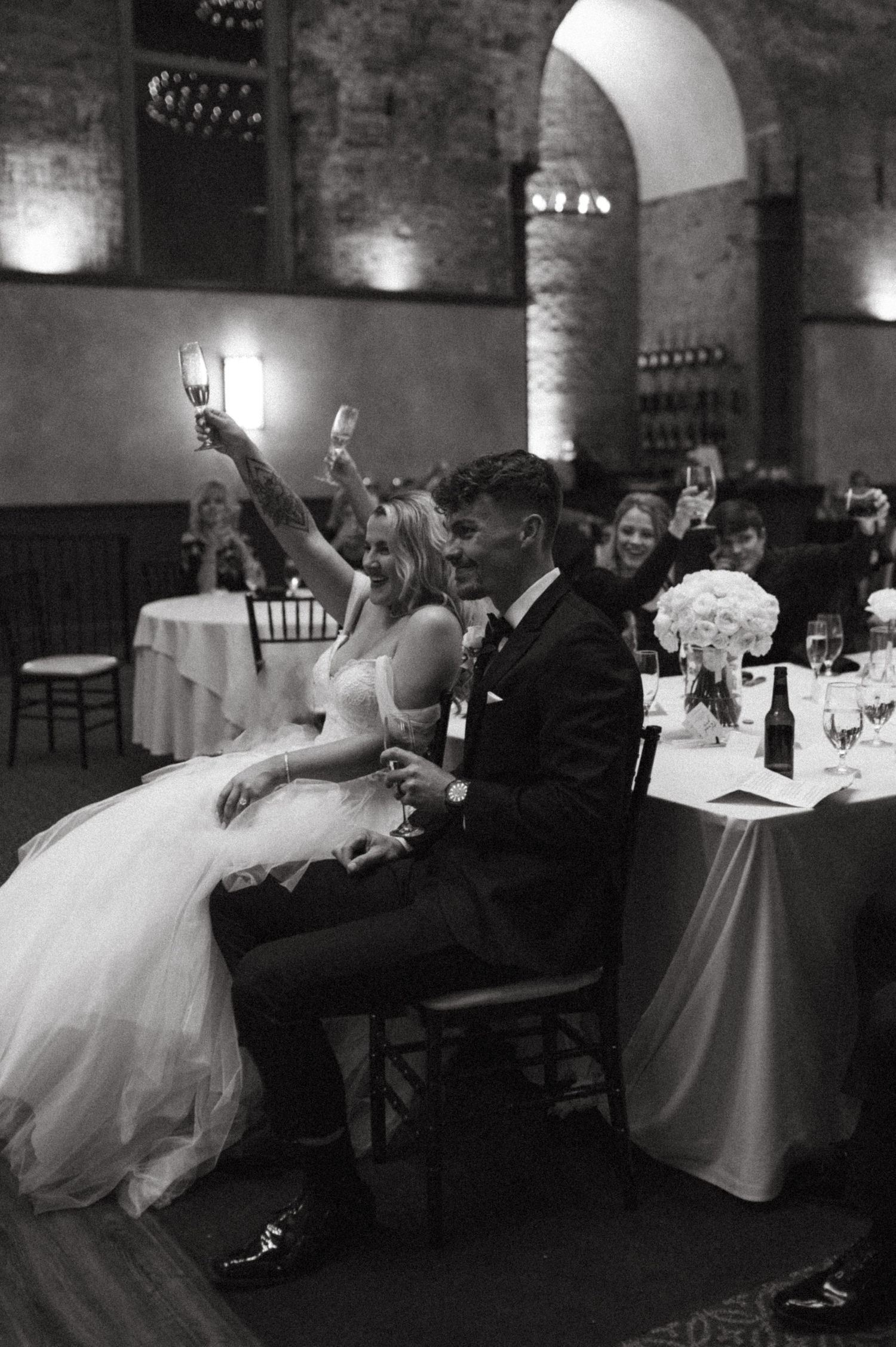 Newlyweds raising champagne glasses during wedding reception toast in historic venue.