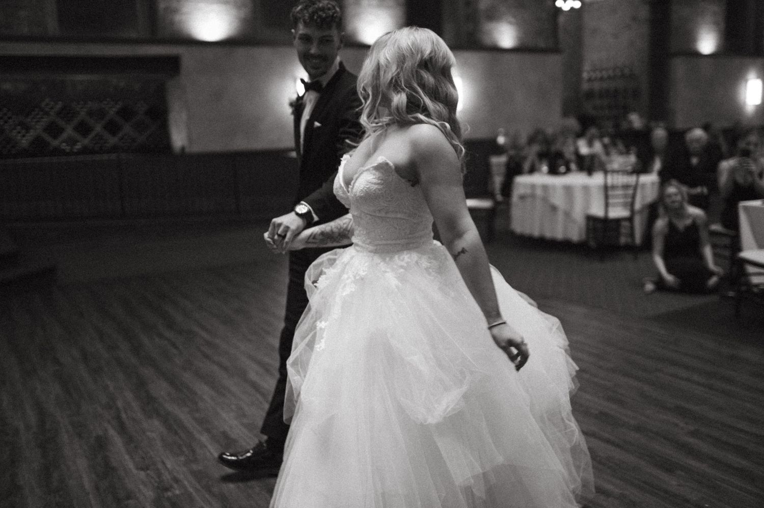 A couple shares their first dance at a wedding reception on a wooden dance floor in black and white.