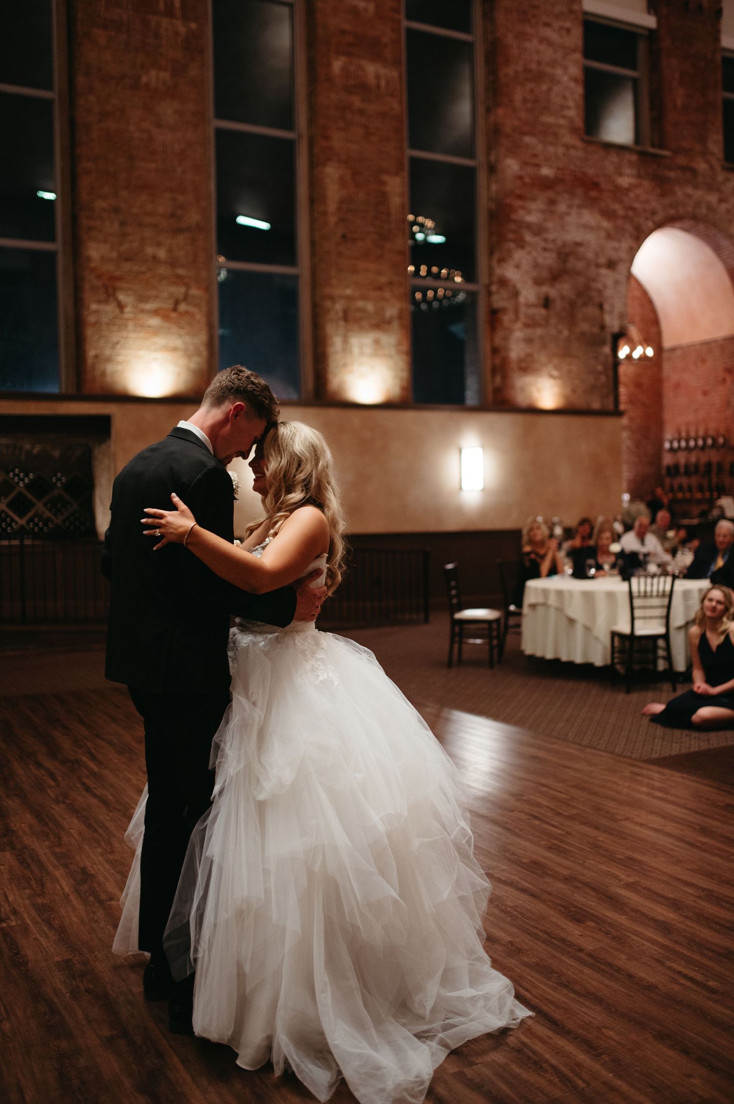 A couple shares an intimate dance moment on a wooden floor with guests watching from decorated tables.