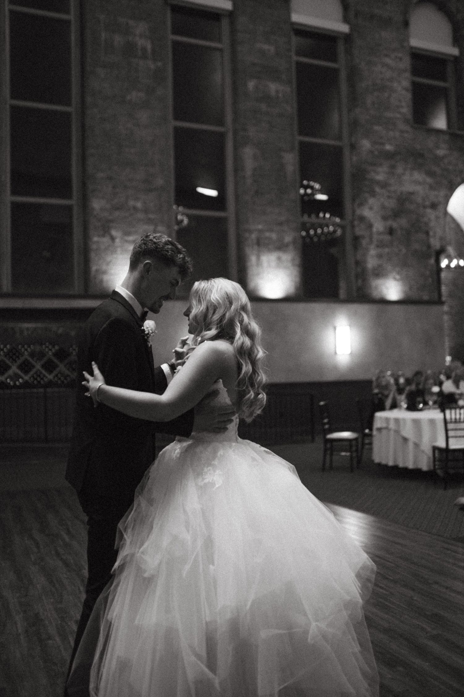 An intimate black and white photo of newlyweds dancing in a rustic venue with exposed brick walls.