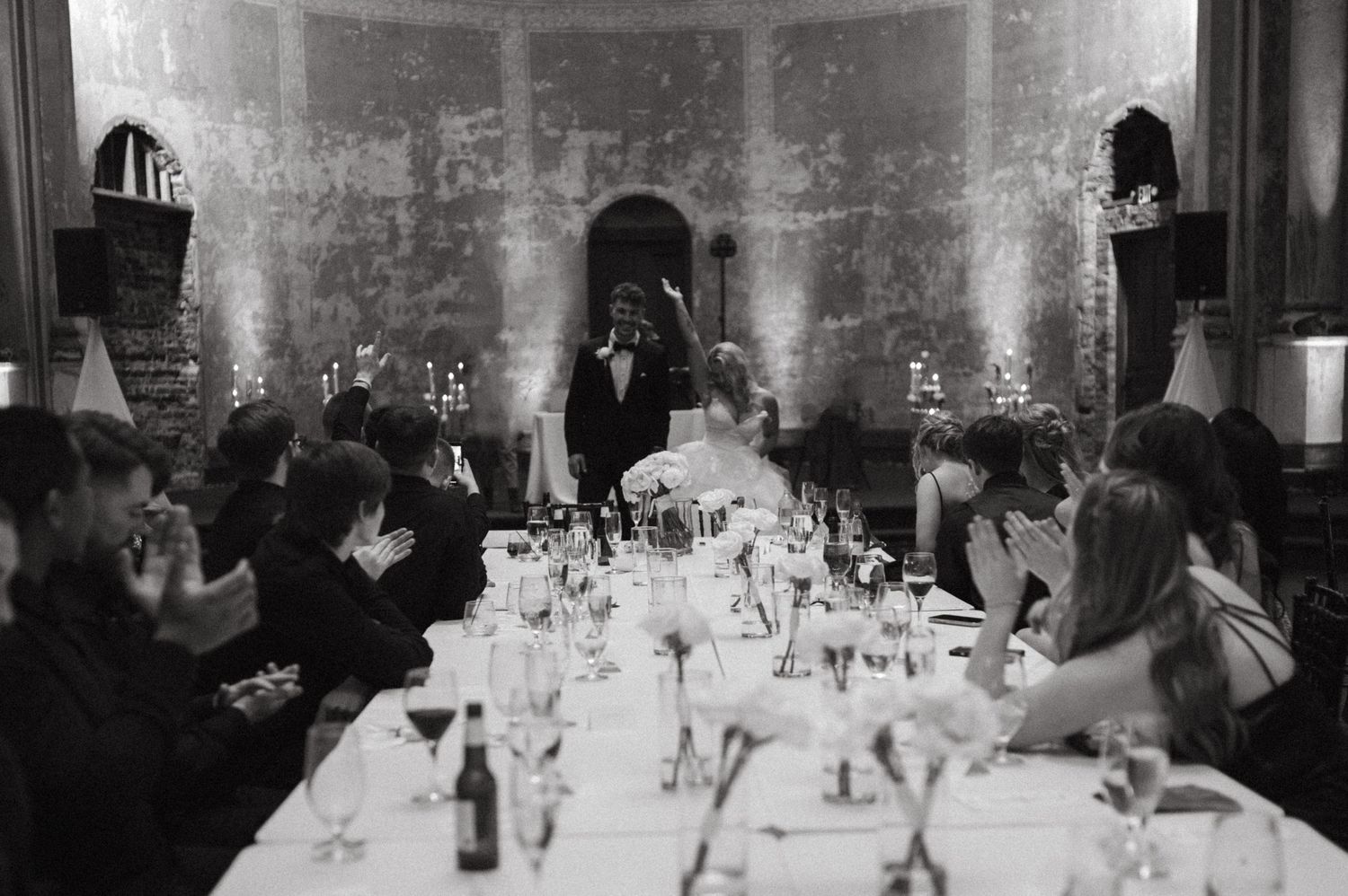 A black and white wedding reception scene with guests seated at long tables in an elegant venue.