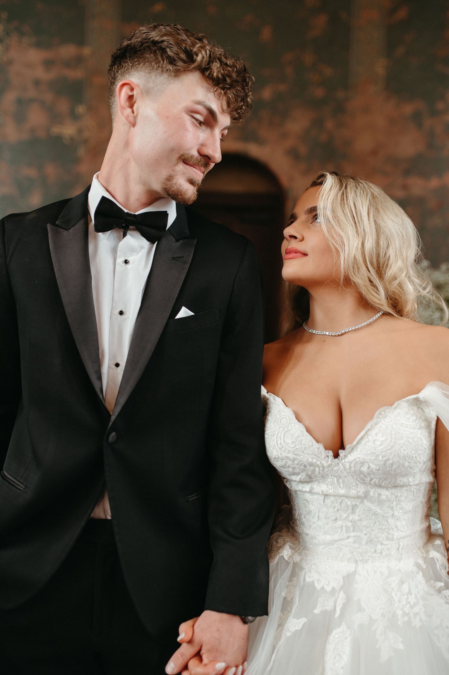 An elegant couple in formal wedding attire pose against a rustic textured wall.