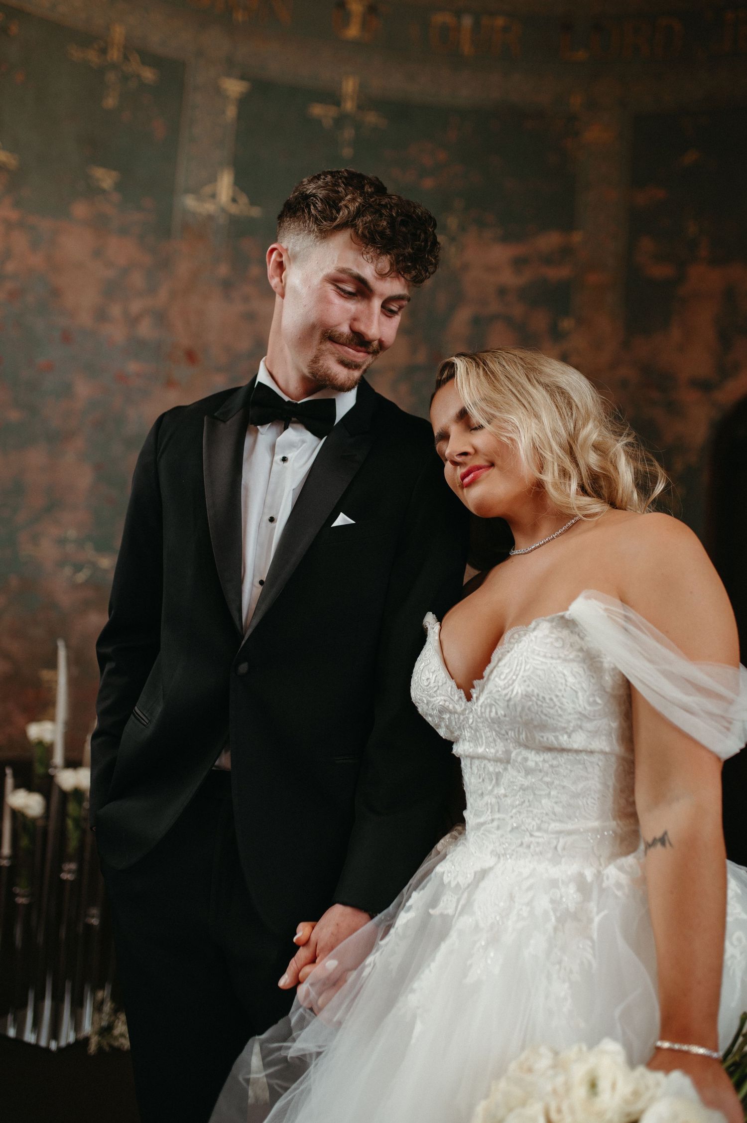 A romantic wedding photo of newlyweds in formal attire against a rustic textured wall with candlelight.