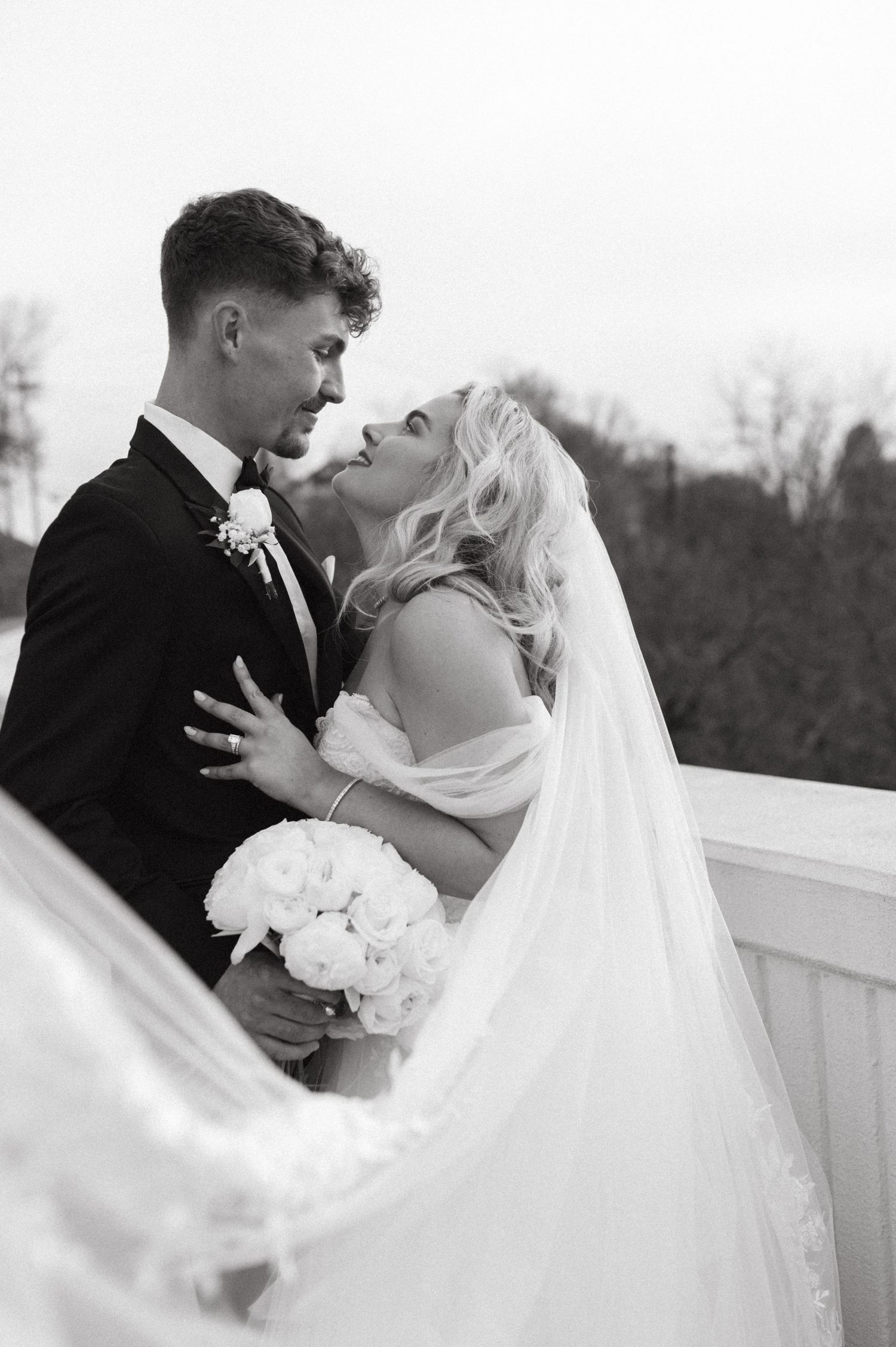 An intimate black and white wedding portrait showing the couple embracing on a balcony with white flowers.