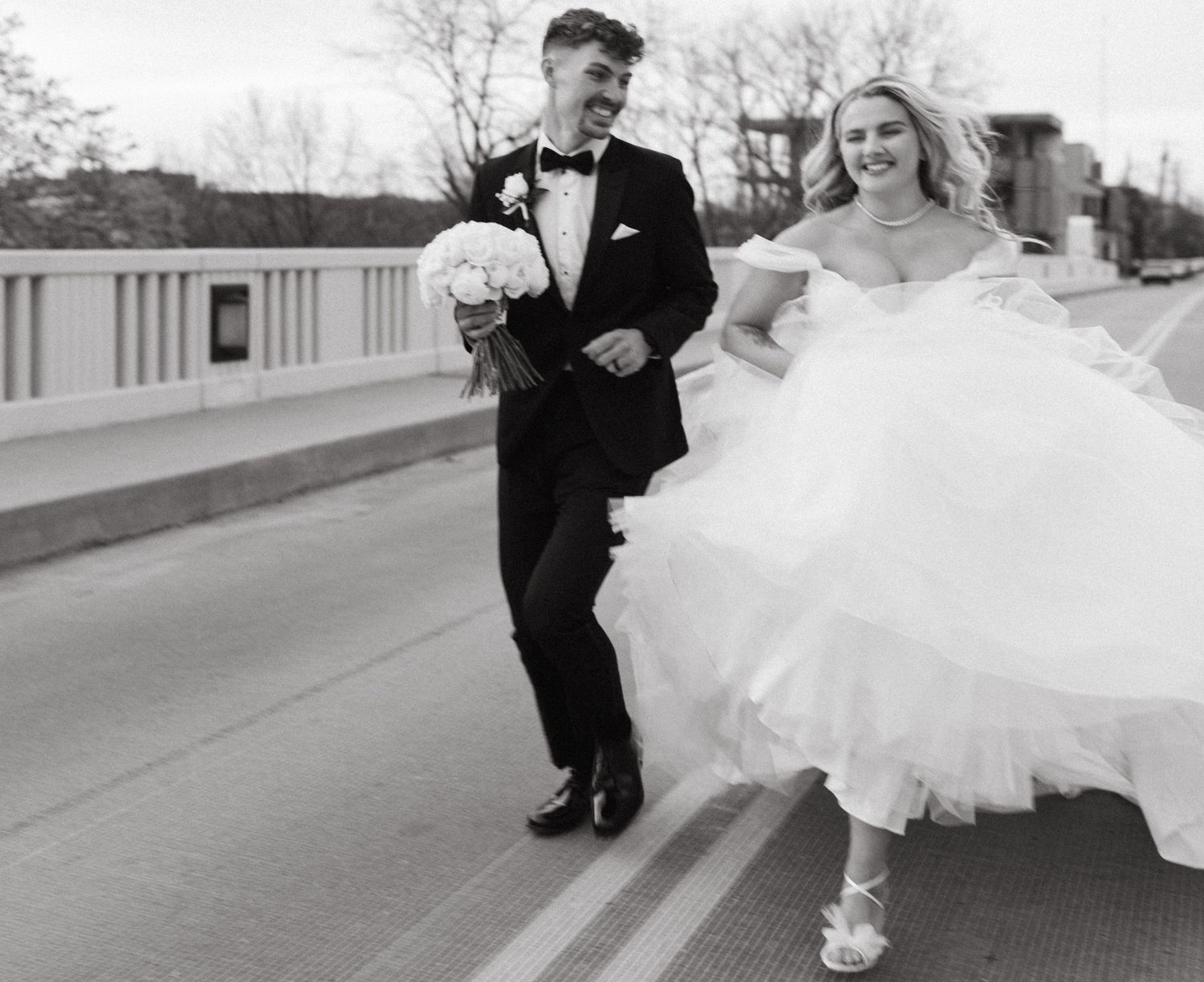 A candid black and white photo of newlyweds running together on a bridge with flowing wedding dress.