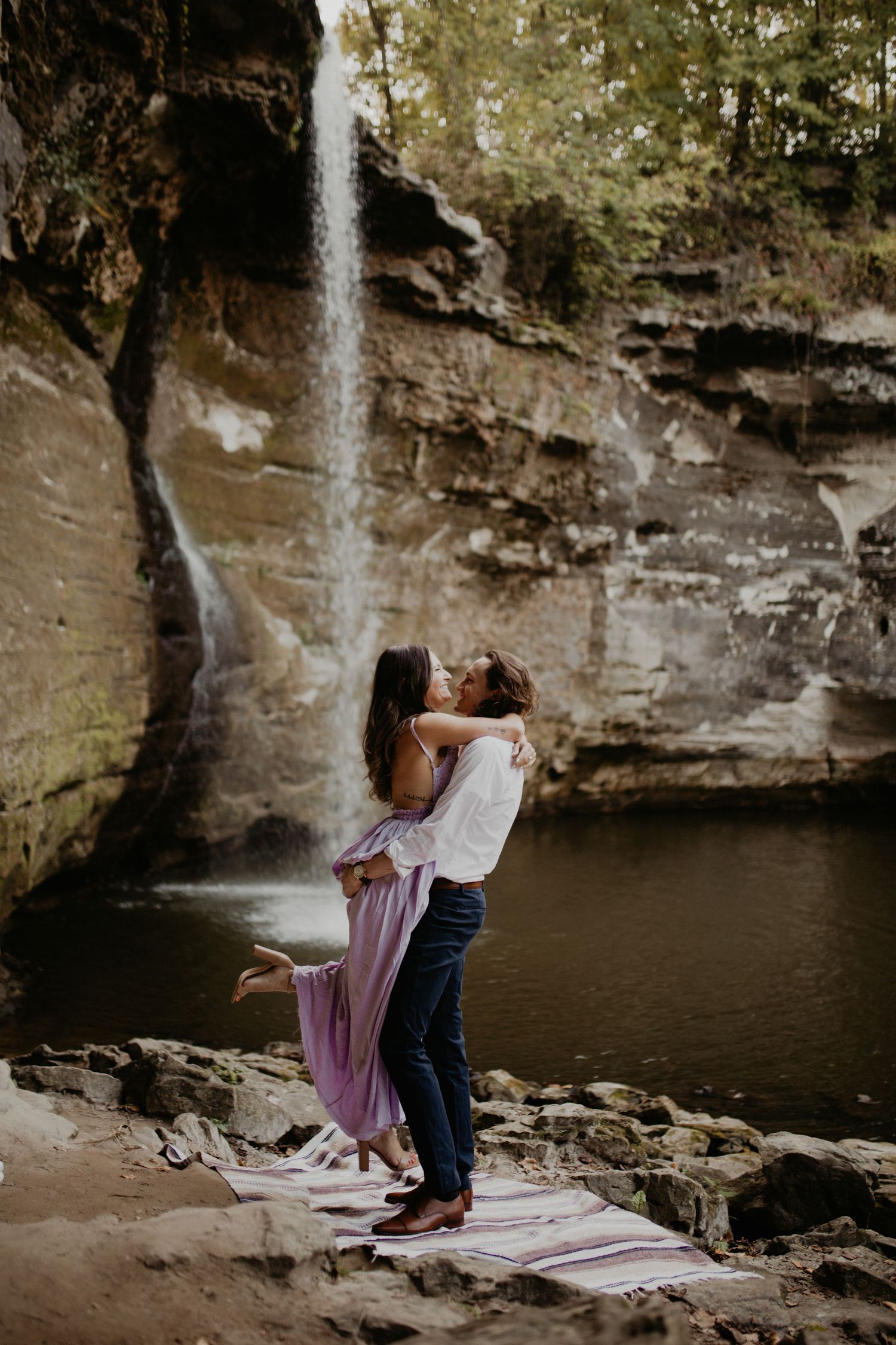 Scenic waterfall cascades down rocky cliffs into a serene pool with figures on a blanket.